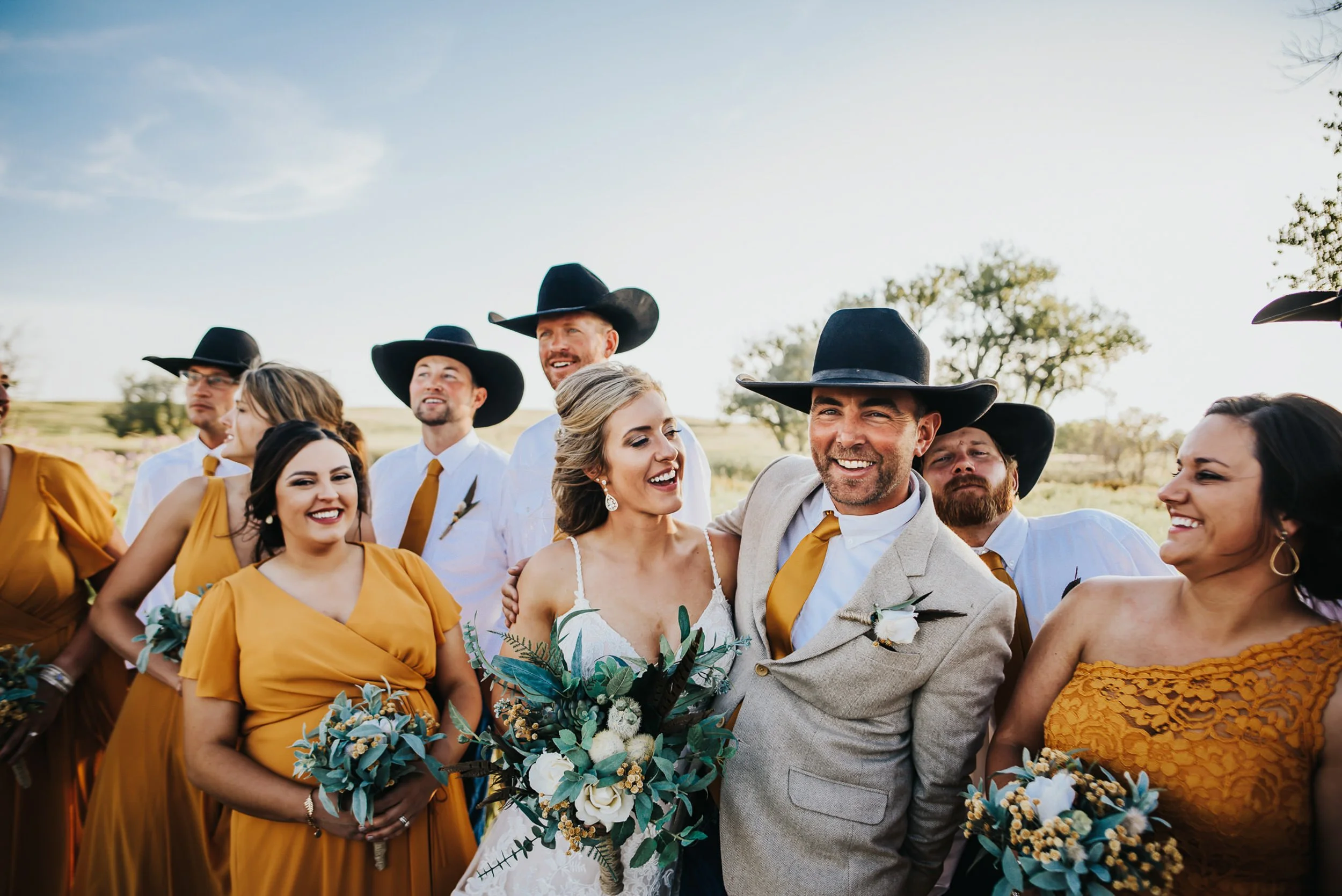 A wedding party in wide-brim hats and mustard and teal tones laughs together outdoors.