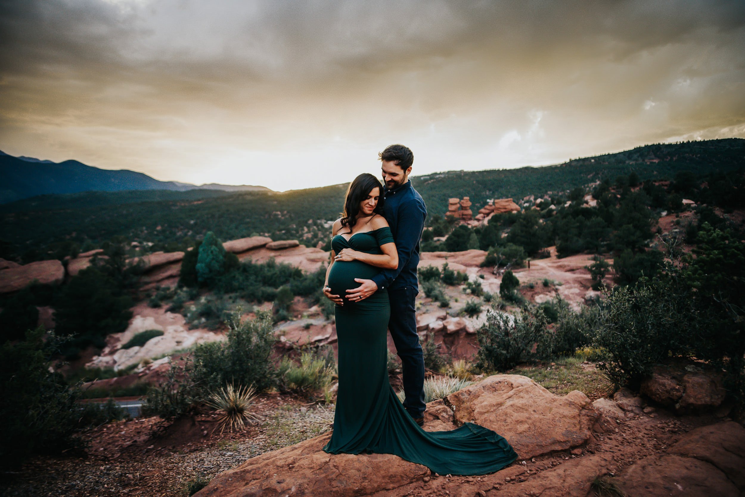 Pregnant couple standing together at Garden of the Gods with red rock formations and a moody sky behind them.