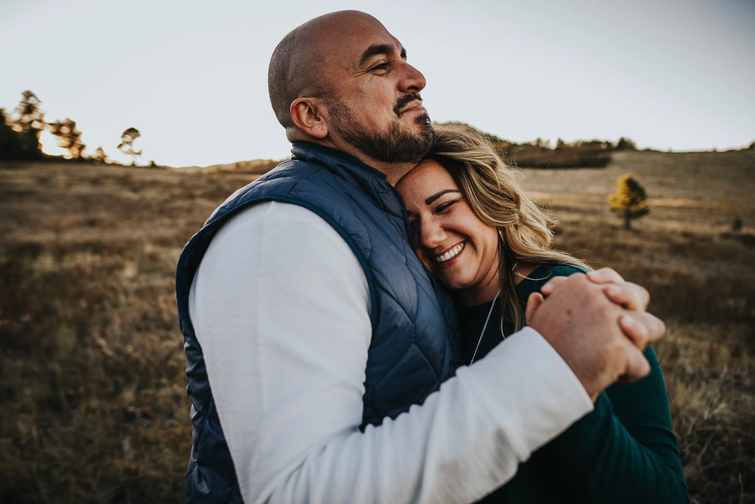A man holds his partner from behind, both laughing warmly in an open Colorado field at Spruce Mountain Open Space.