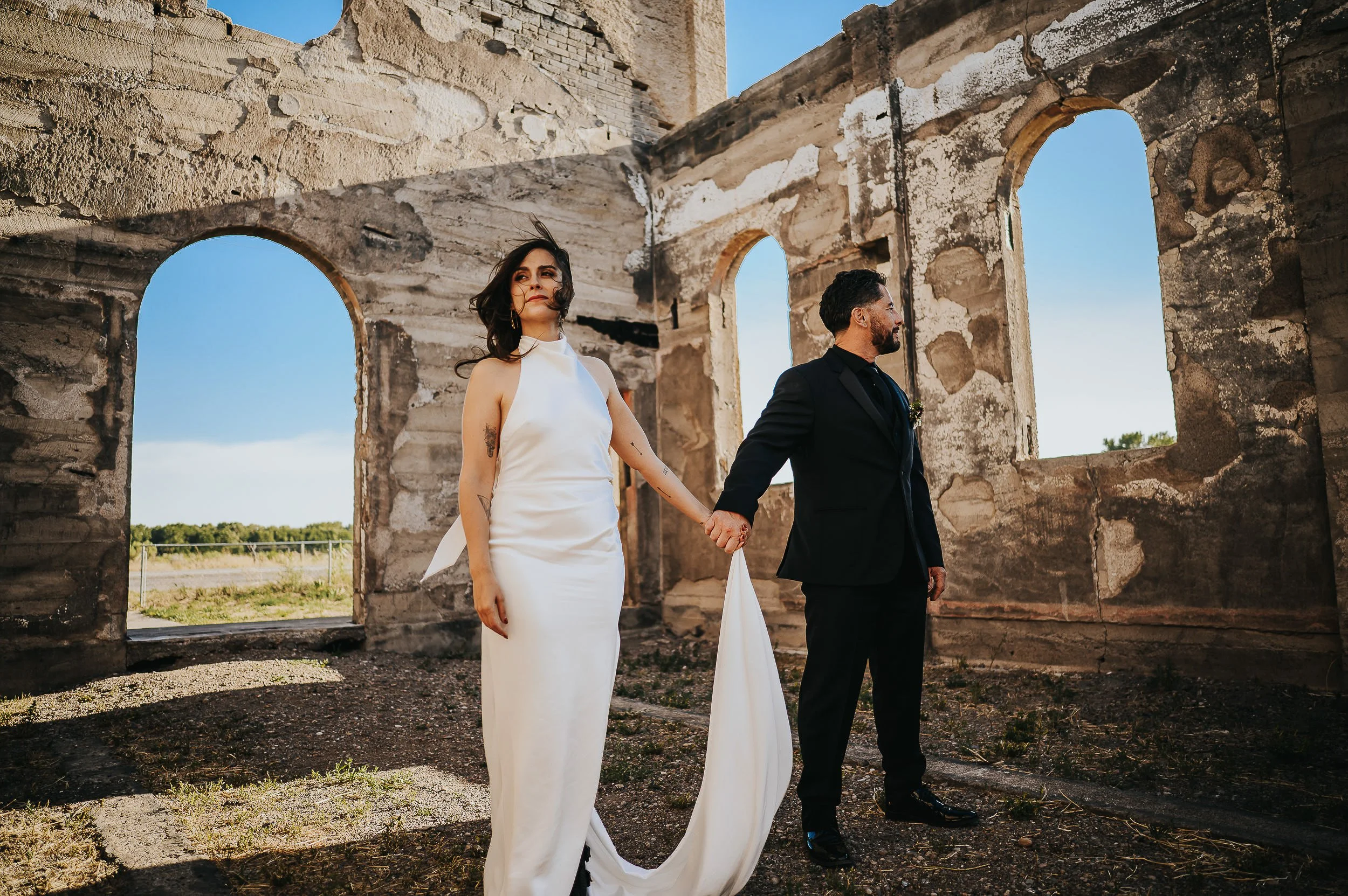 Bride and groom standing hand in hand inside an open stone ruin with arched windows and mountain views.