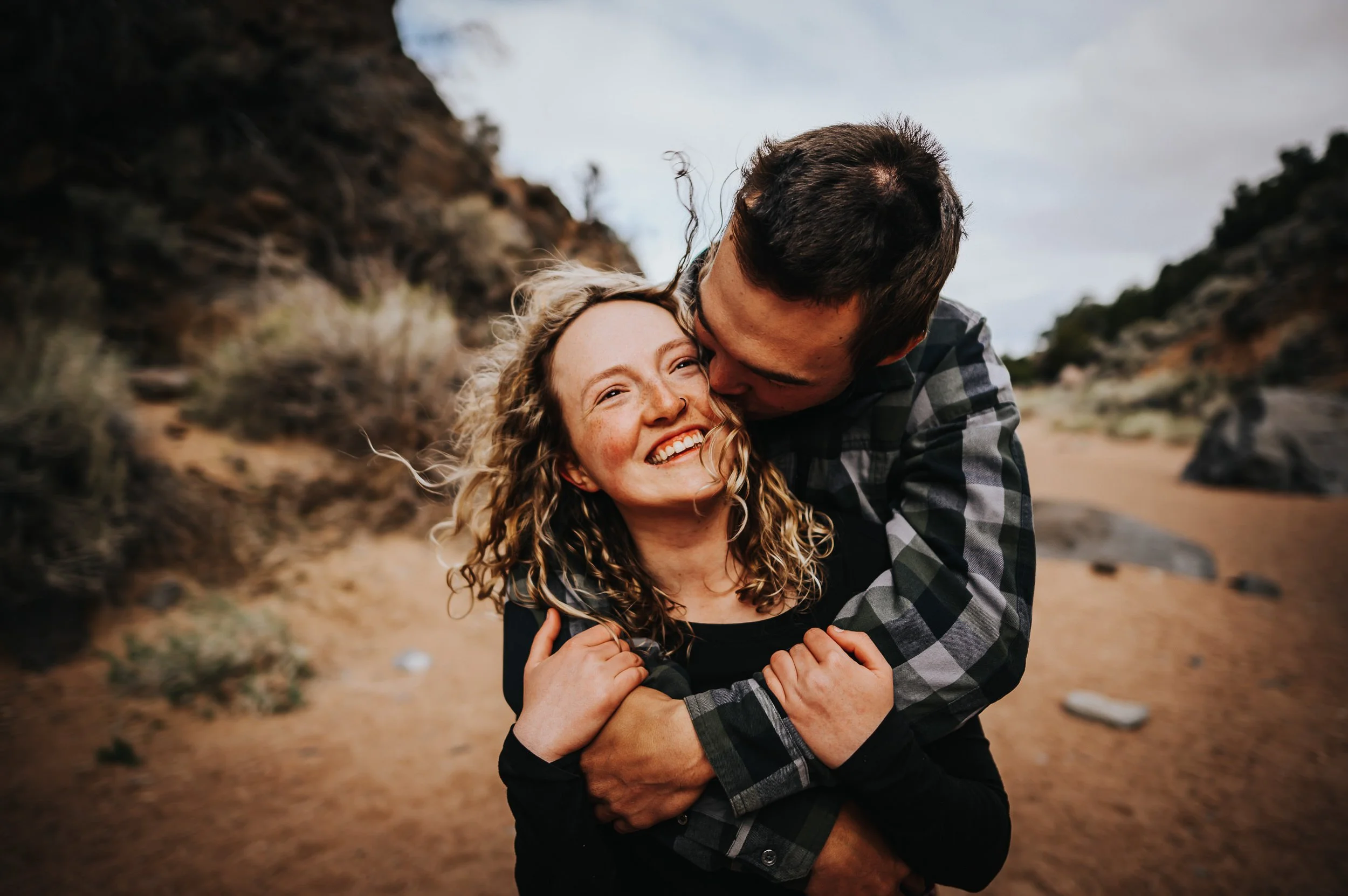 A woman laughs as her partner wraps around her from behind on a Colorado trail, both in casual layers.