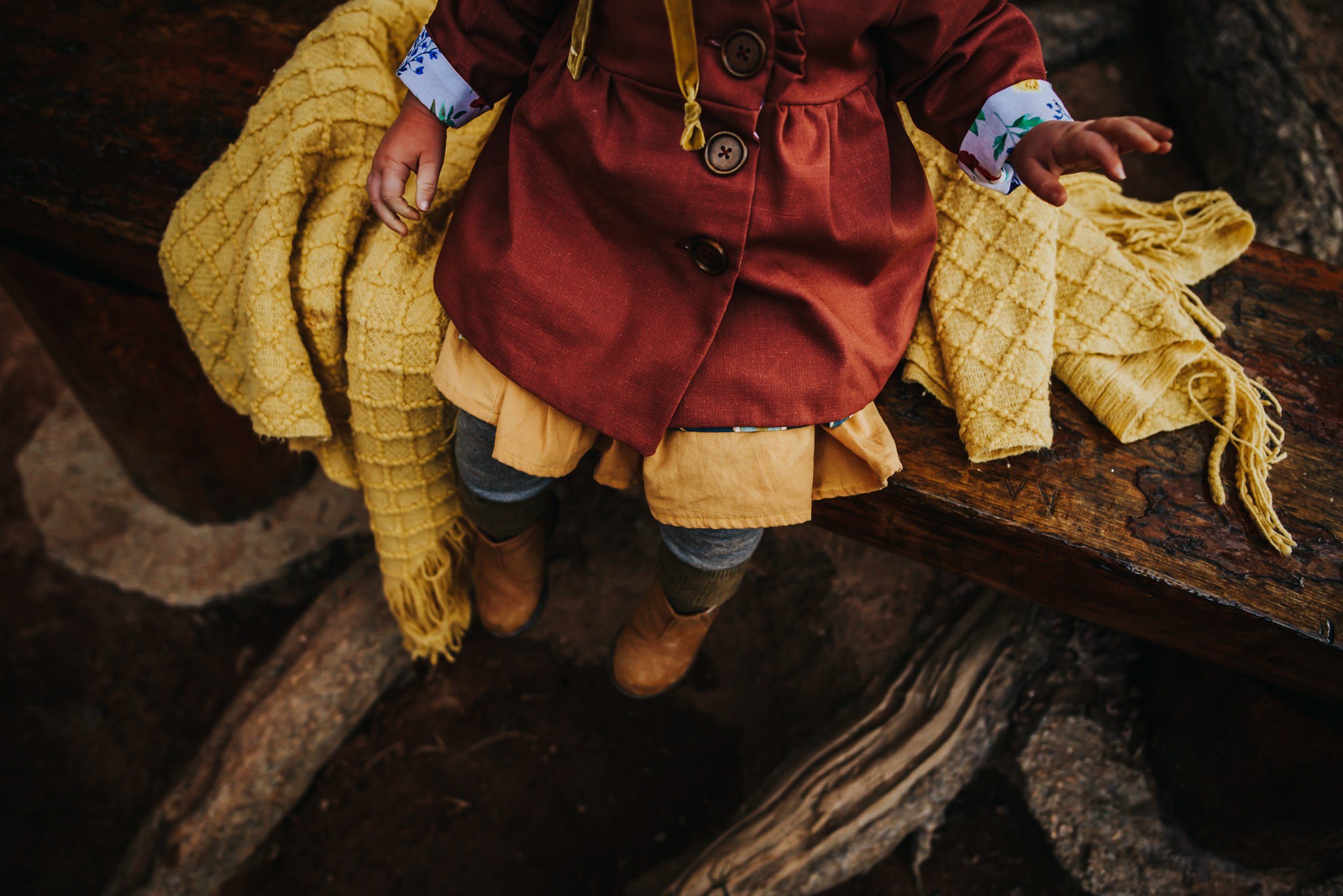 A child sitting on a yellow blanket on a natural wooden bench wearing a red coat photographed from overhead.