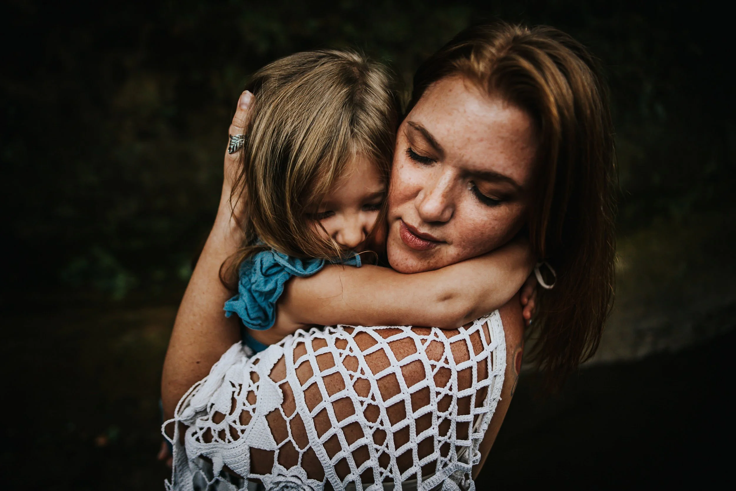 Mom in cream crochet top embracing two daughters, one in teal, by a rocky Colorado creek.