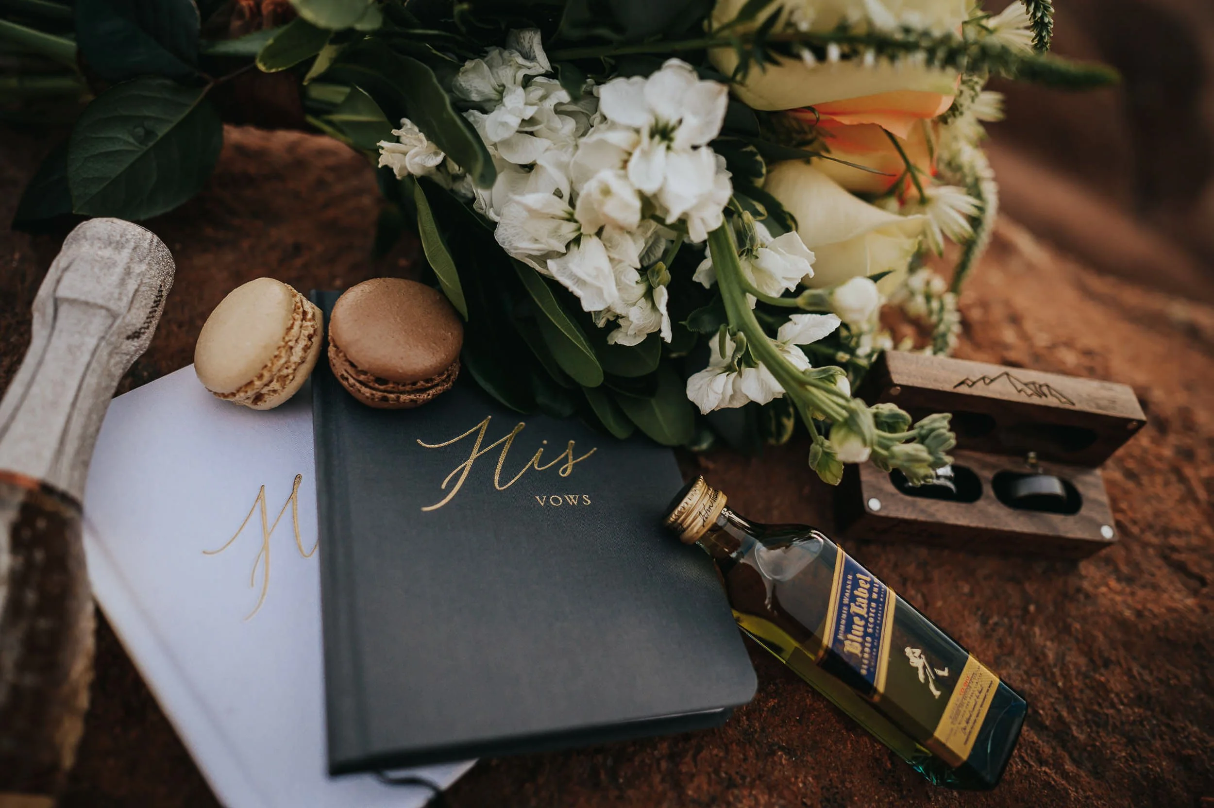 A wedding flatlay of white florals, macarons, a vow book, and rings on a dark surface.