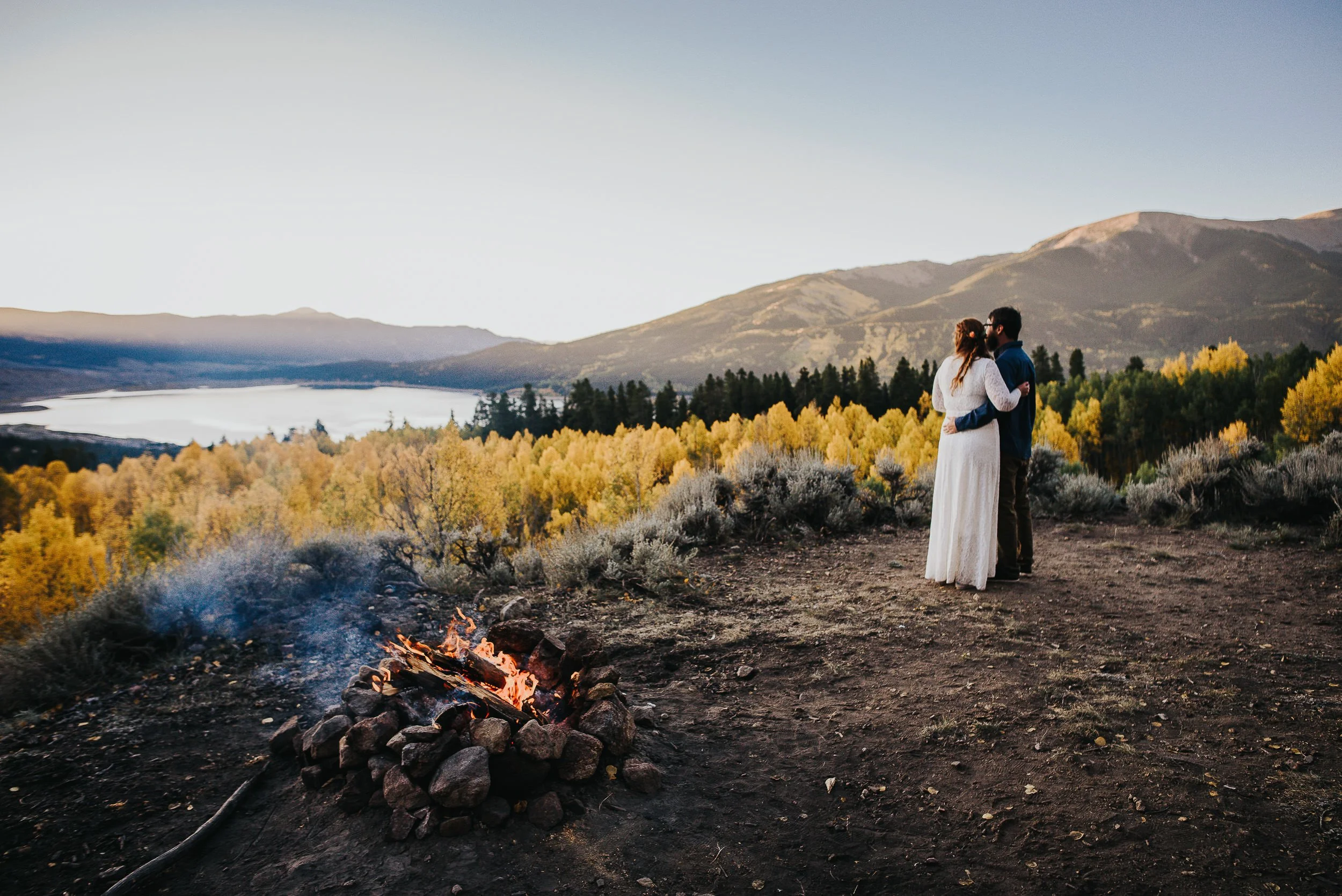 A couple stands beside a campfire at the edge of an alpine lake, fall aspens glowing behind them.