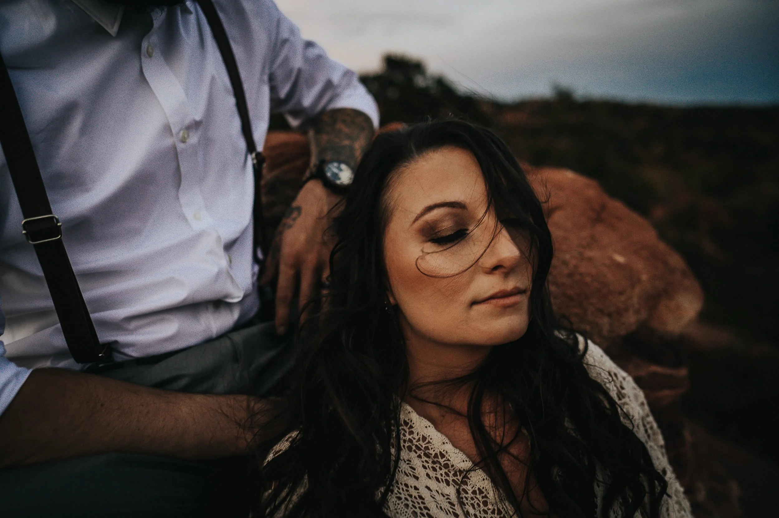 A woman in a lace top leans back with eyes closed in a moody closeup portrait outdoors.