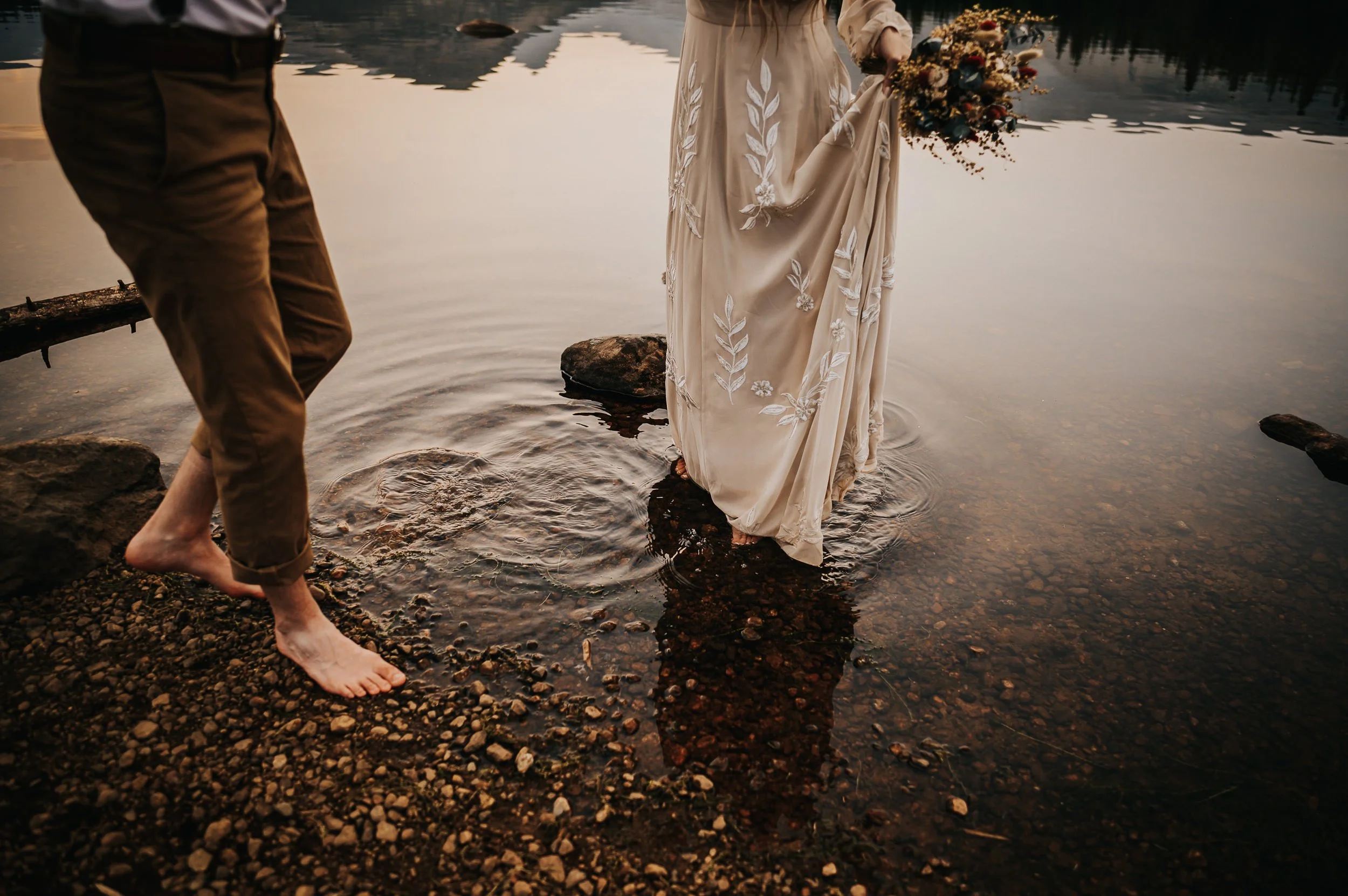 A bride stands barefoot in shallow water, her lace dress and bouquet reflected in the surface.