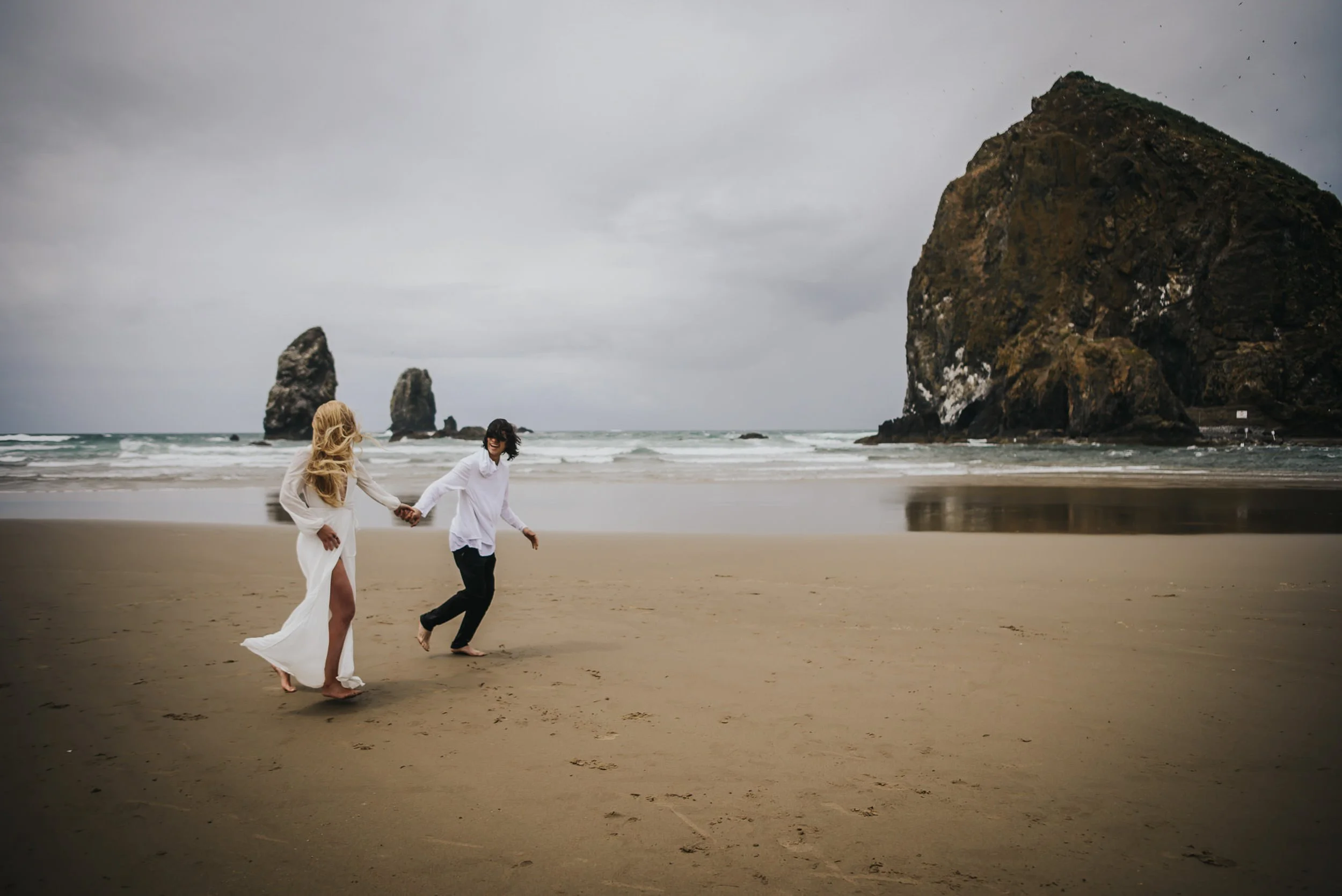 A couple runs hand in hand down a wide sandy beach, sea stacks visible in the distance.