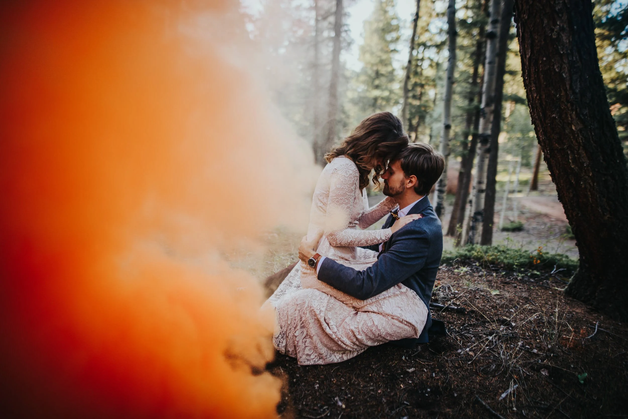 A couple sits wrapped together in orange smoke bomb haze in a Colorado forest.