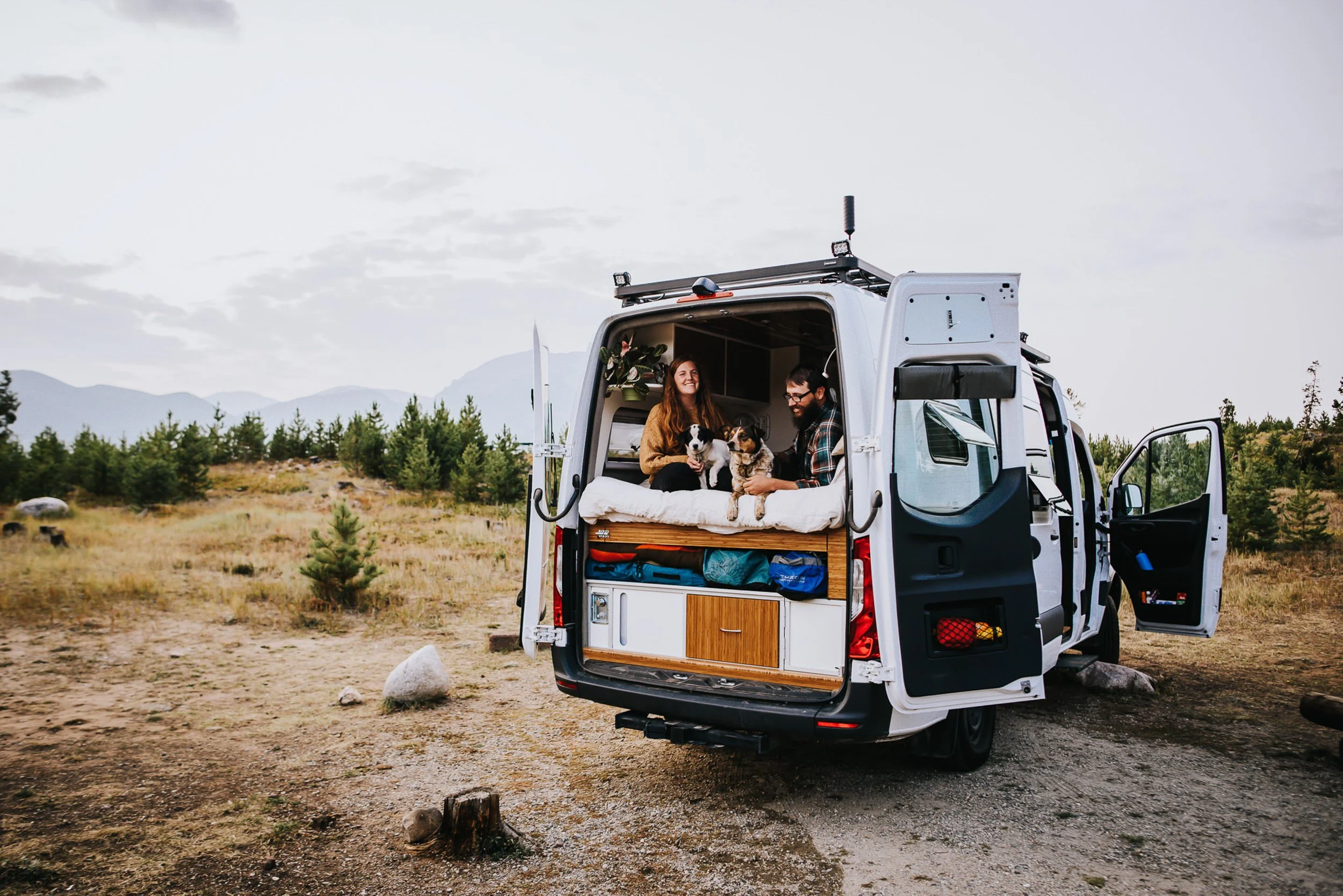 A couple stands in the open back of a camper van parked in a Colorado forest, relaxed and free in Frisco, Colorado.