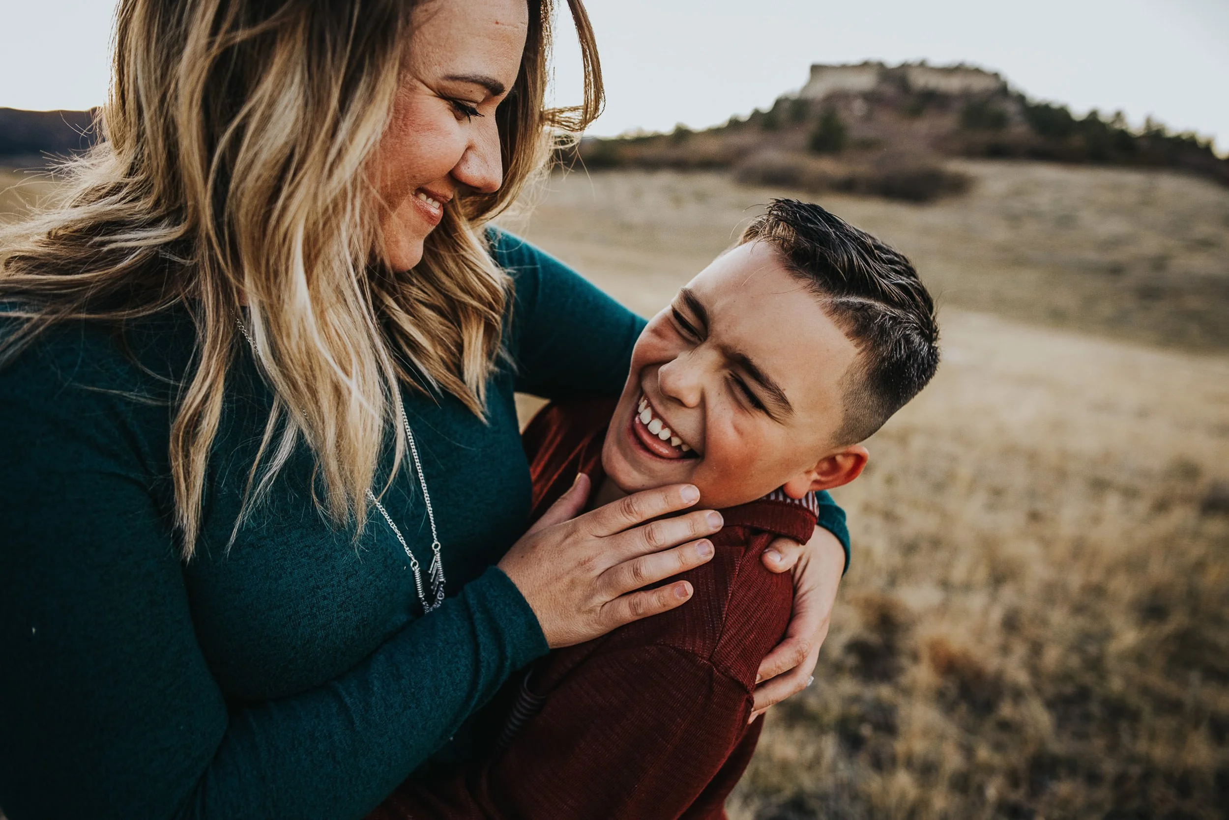 A mom and son press cheek to cheek laughing outdoors, warm and joyful closeup.