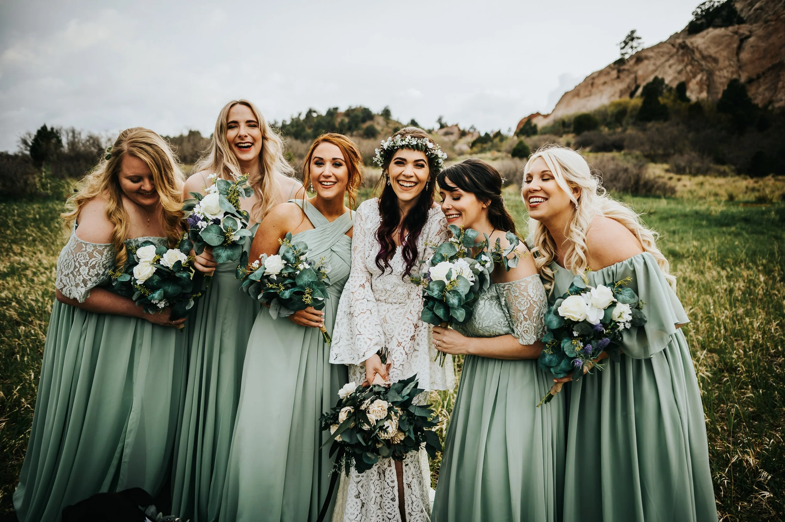 A bride with a flower crown laughs with her bridesmaids in sage green dresses at Garden of the Gods, red rocks behind them.
