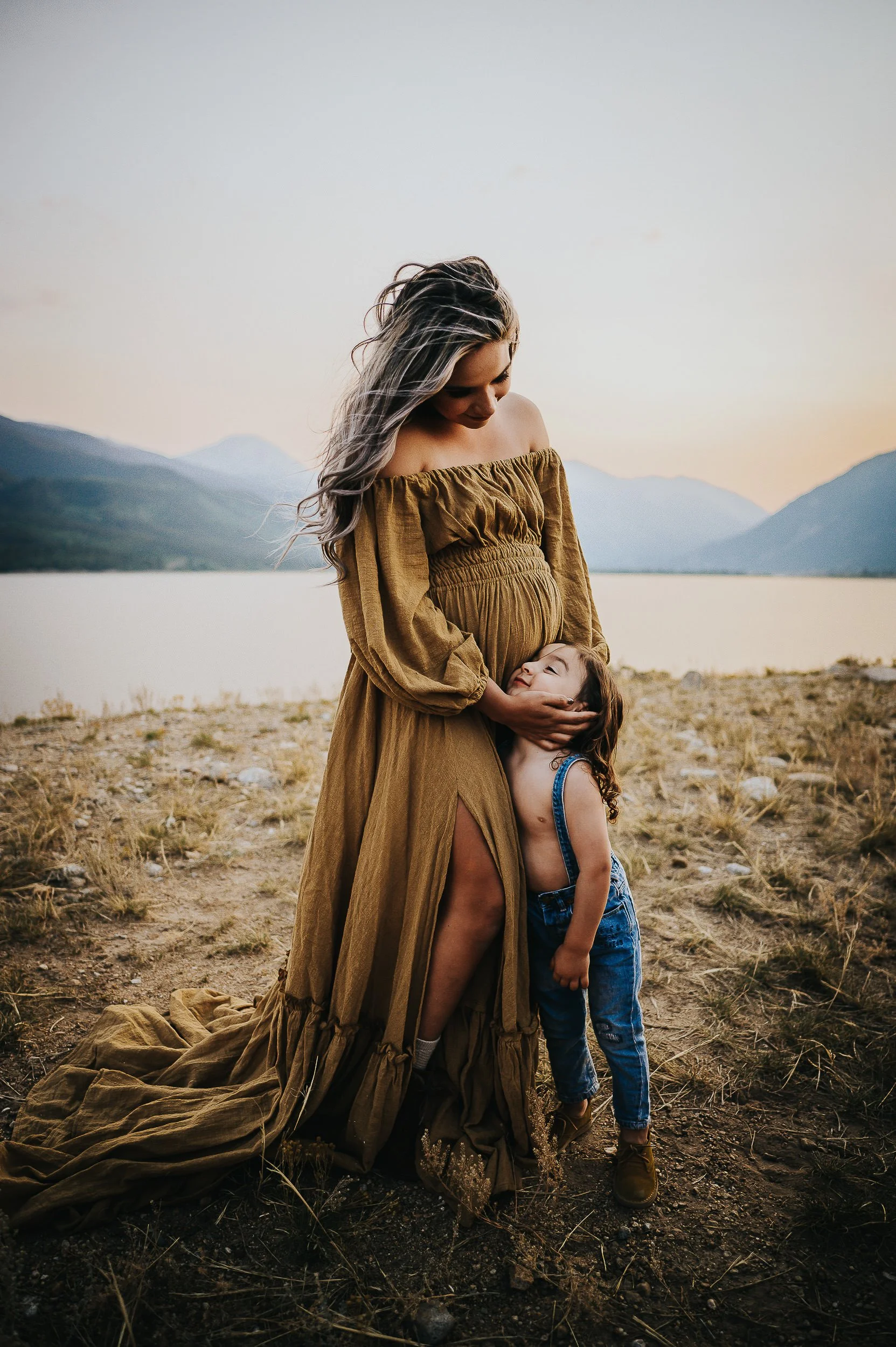 Pregnant woman in an olive dress with a young child hugging her belly beside a mountain lake.