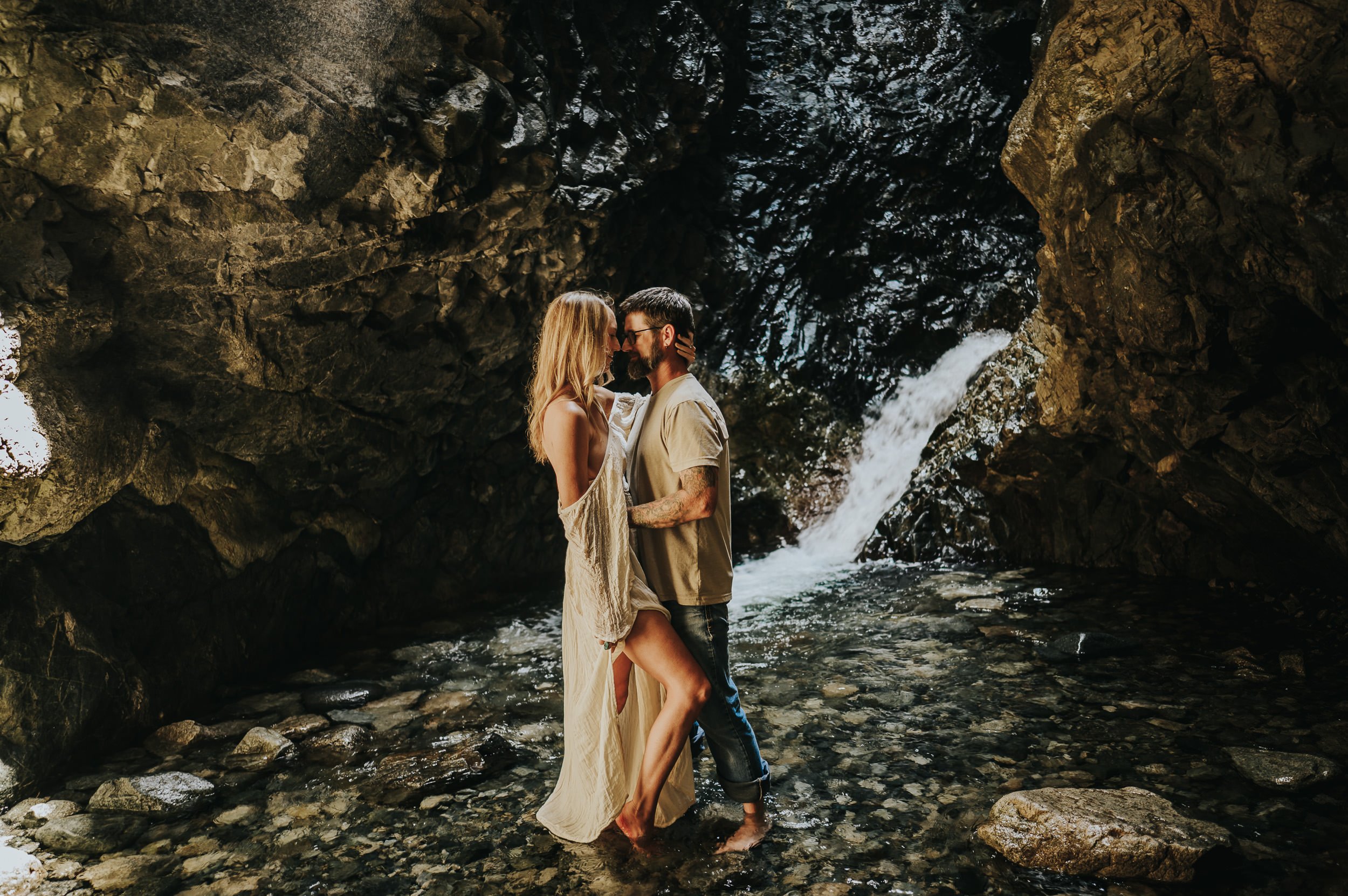 A couple embraces each other in front of a waterfall while wearing neutral cream and ivory tones.