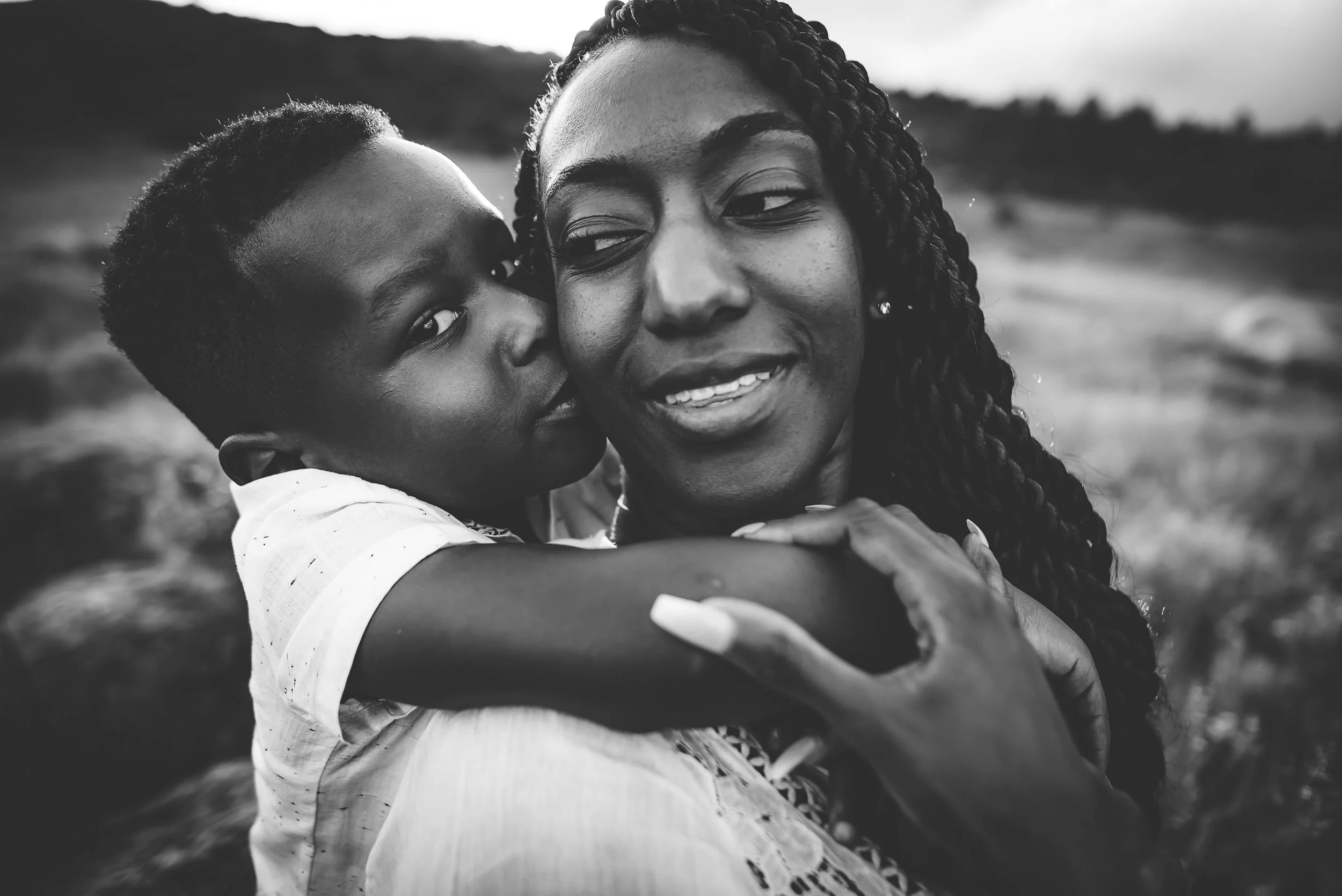 A mom and son embraces tightly, faces close, in a black and white grassy field portrait.