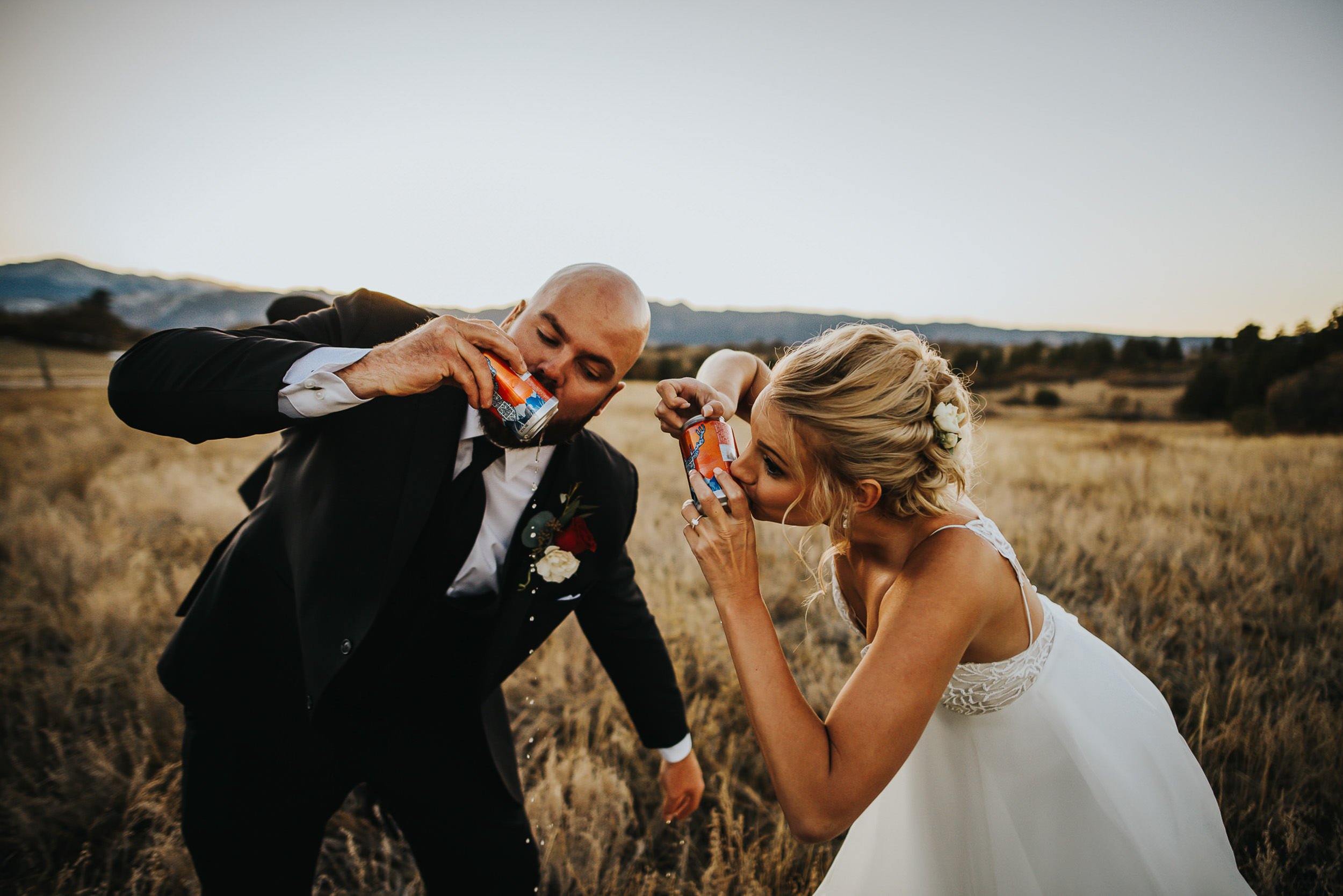 A groom in a black suit drinks beer with his bride in a field, she in an updo and white gown, both laughing softly.