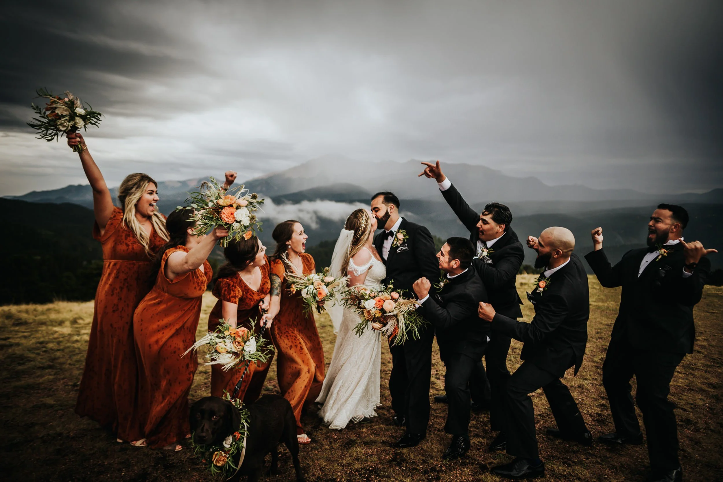 A wedding party celebrates together on a hilltop, everyone raising bouquets and arms in the air.