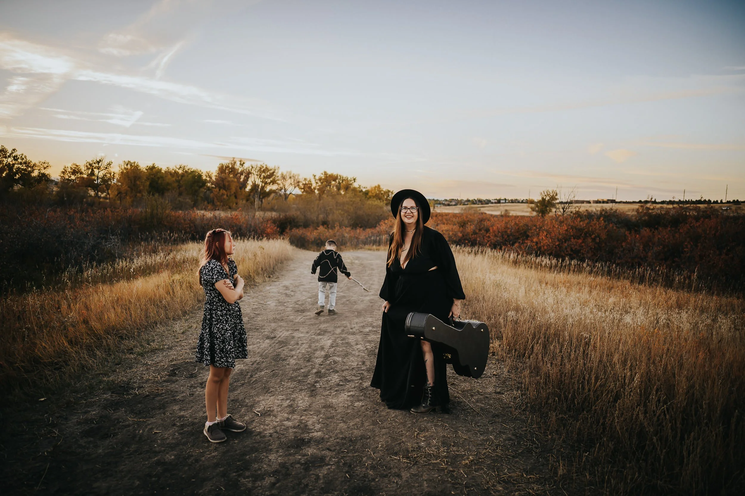A mother and her children play on a hiking trail surrounded by tall golden grasses while wearing a black dress, black hat, and carrying a guitar case.