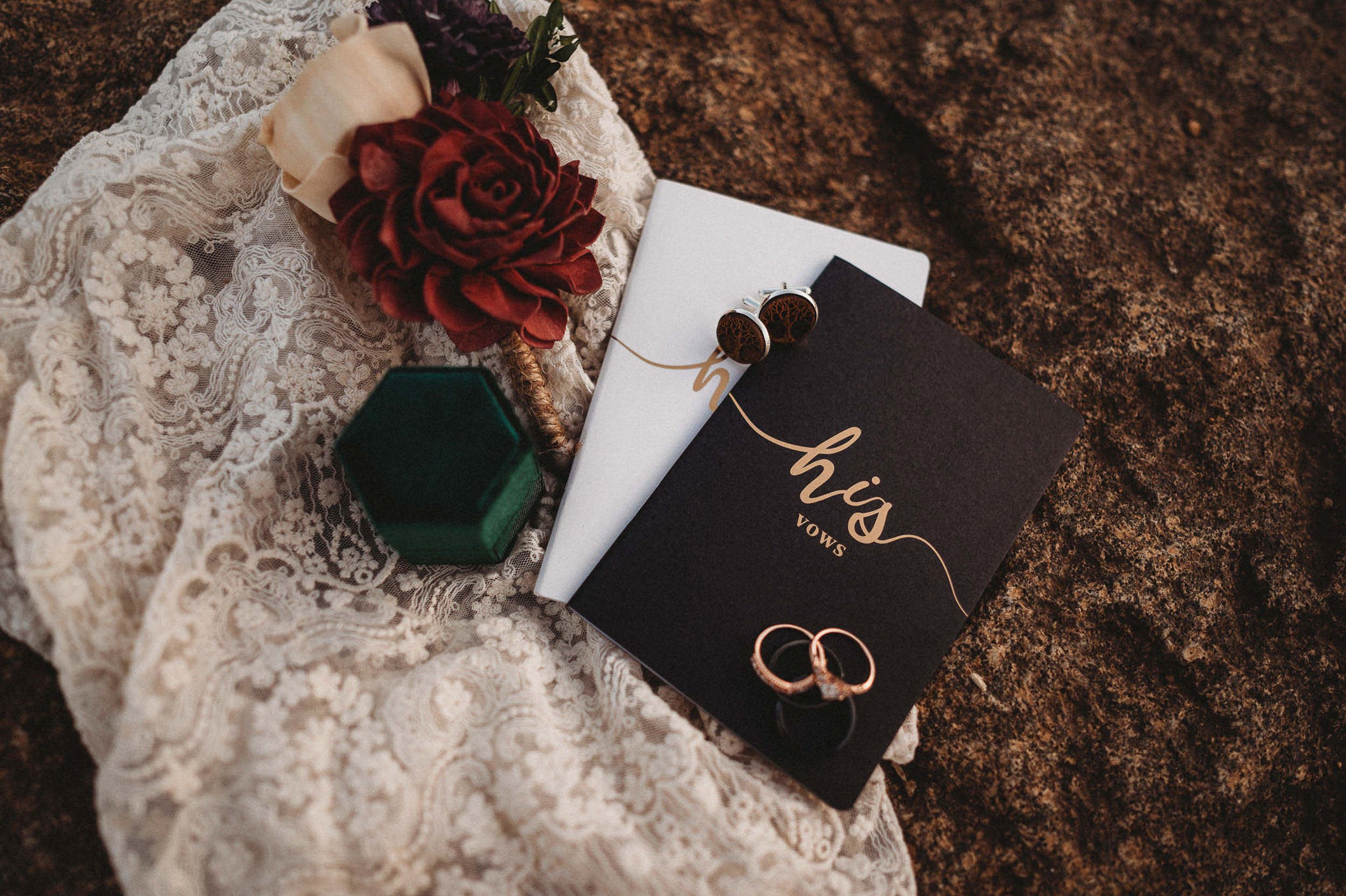 A wedding flatlay with rings, a burgundy floral, and a vow booklet on a rocky surface.