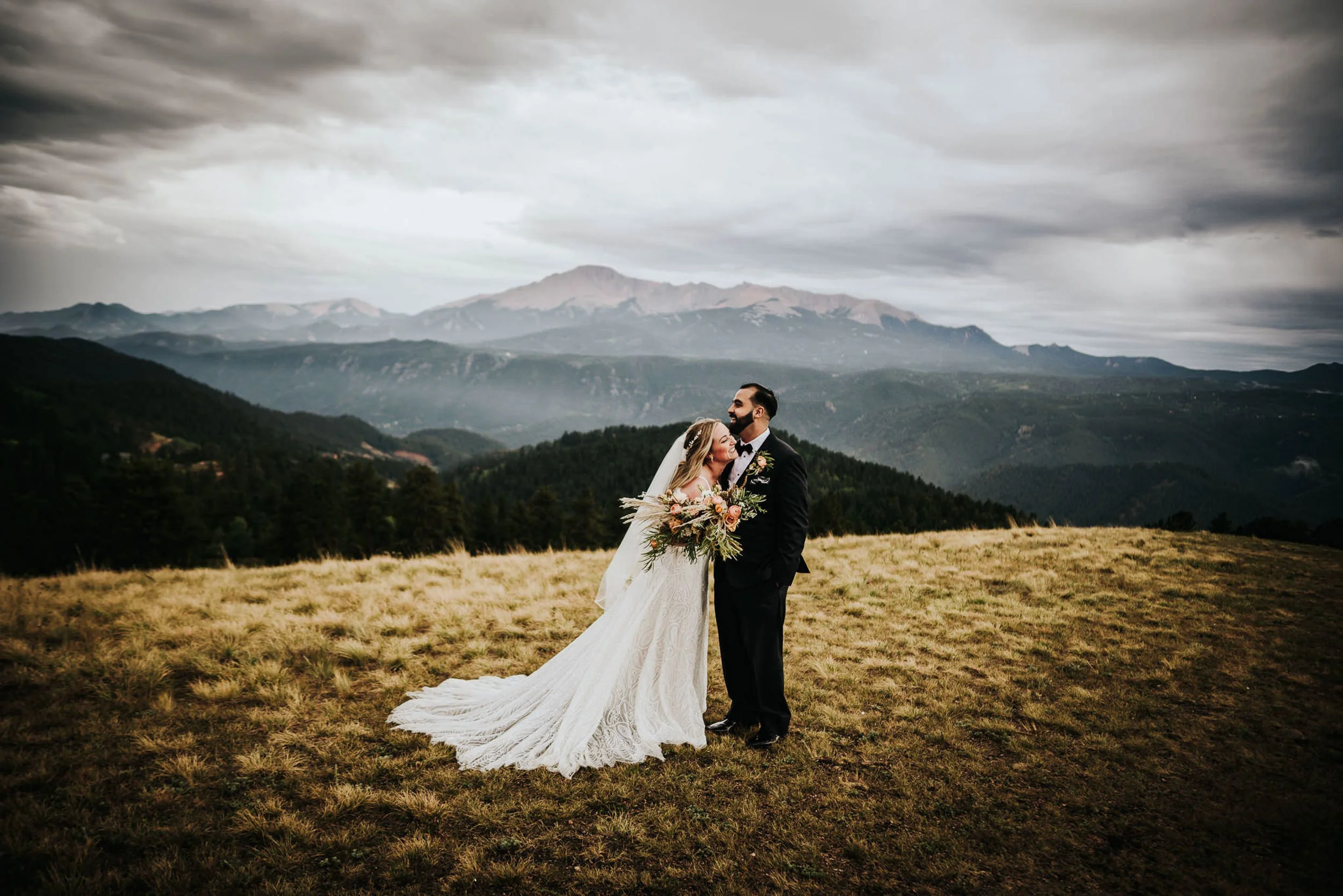 A bride in a full lace gown stands with her groom in a wide Colorado mountain meadow, peaks stretching behind them.