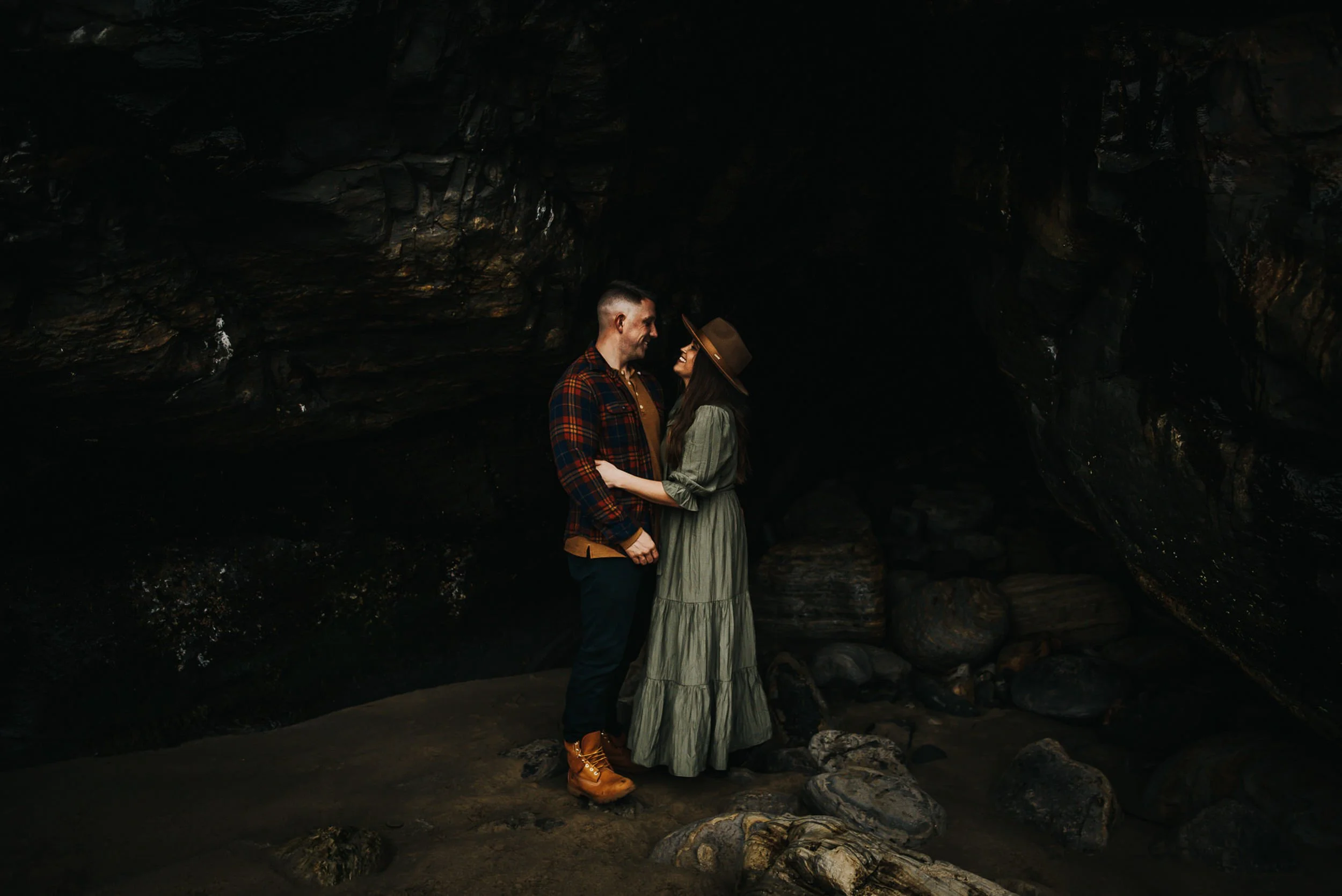 A couple stands close together in a shadowy forest clearing, she in a sage green dress, warm and quiet.