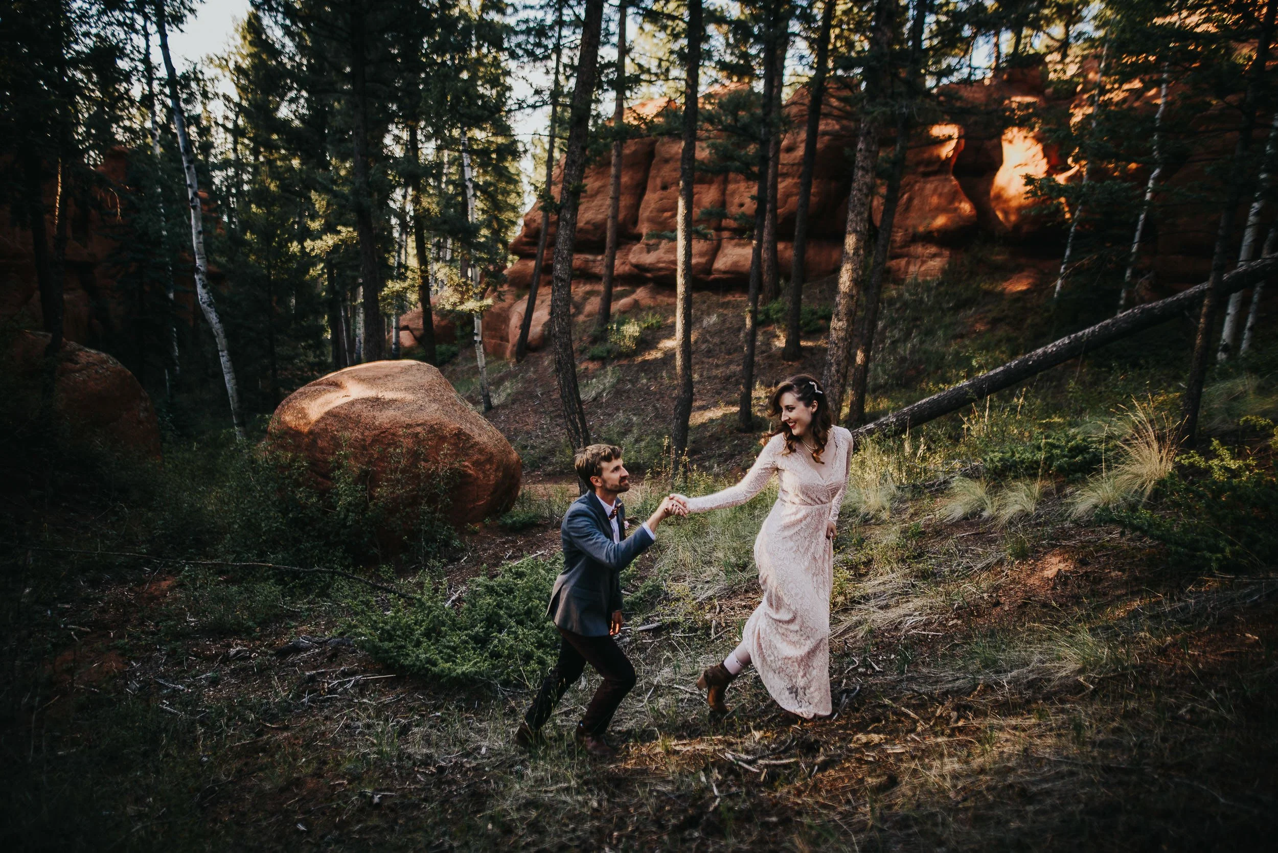A bride runs through a sunlit Colorado forest, groom chasing after her, both laughing in Woodland Park, Colorado.