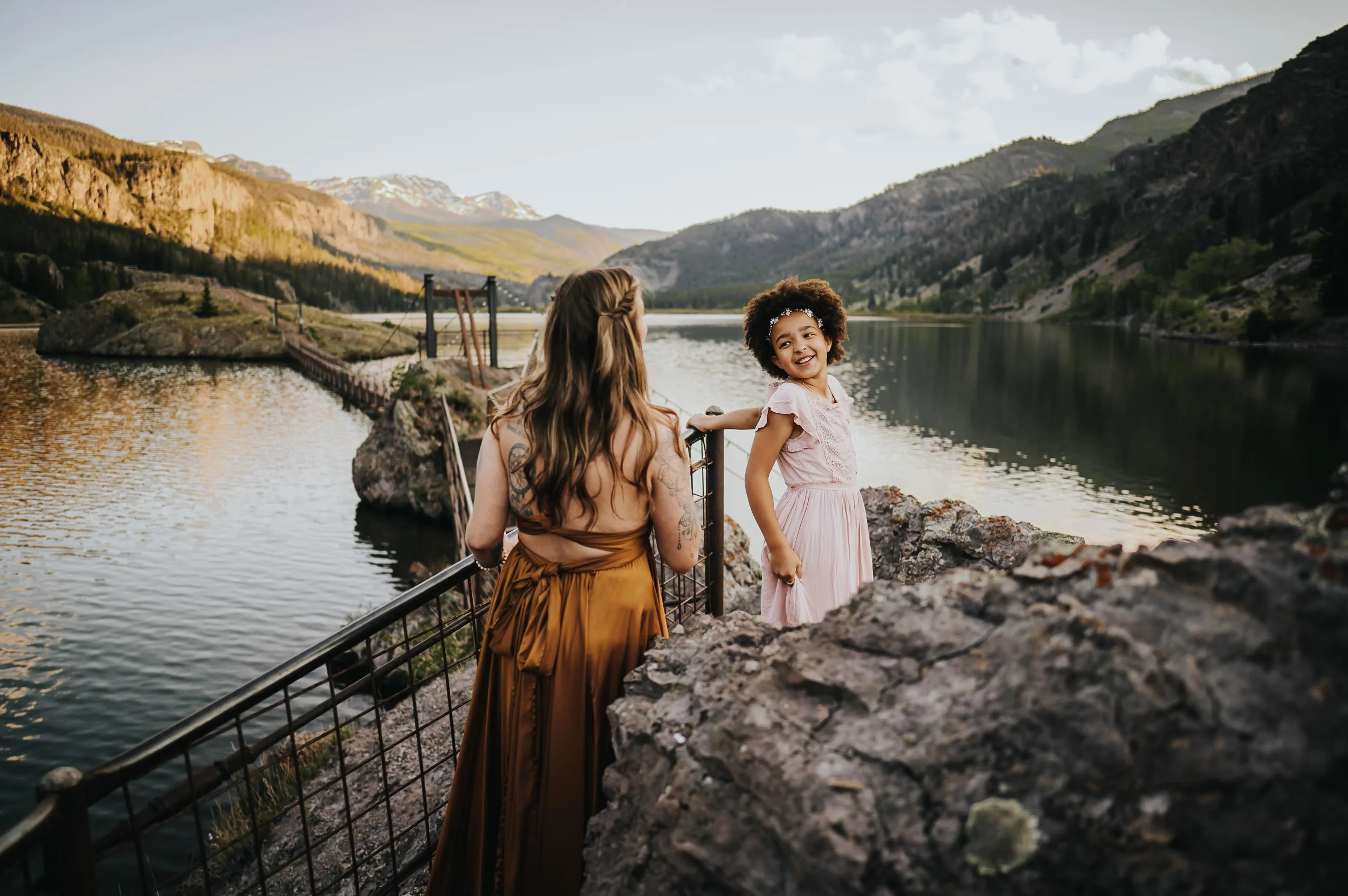 Family of two at an alpine lake with mountains behind them, daughter in mauve dress, natural connected moment.