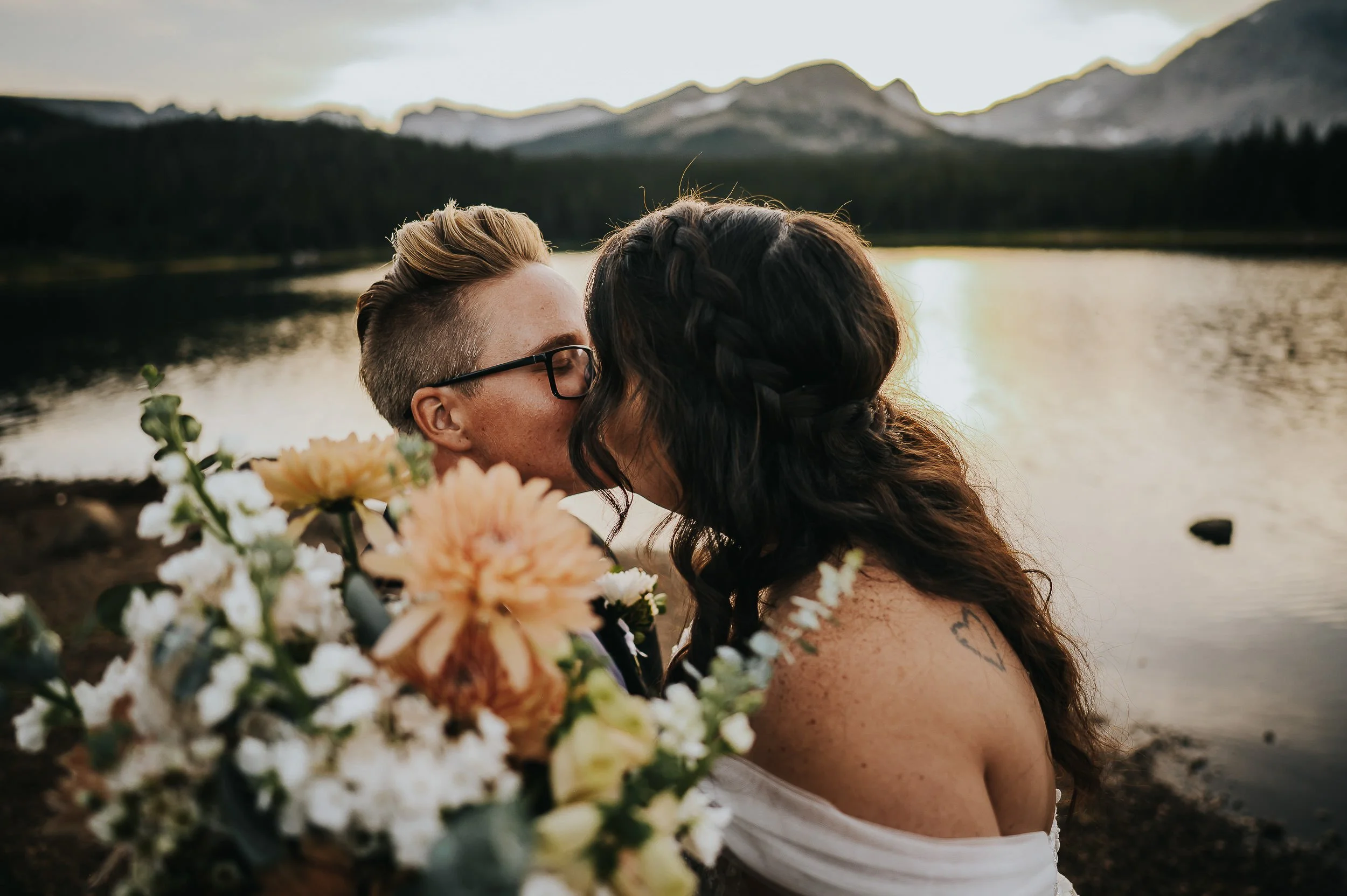 A couple shares a forehead-to-forehead kiss in front of a Colorado mountain lake, her bouquet of dahlias between them.