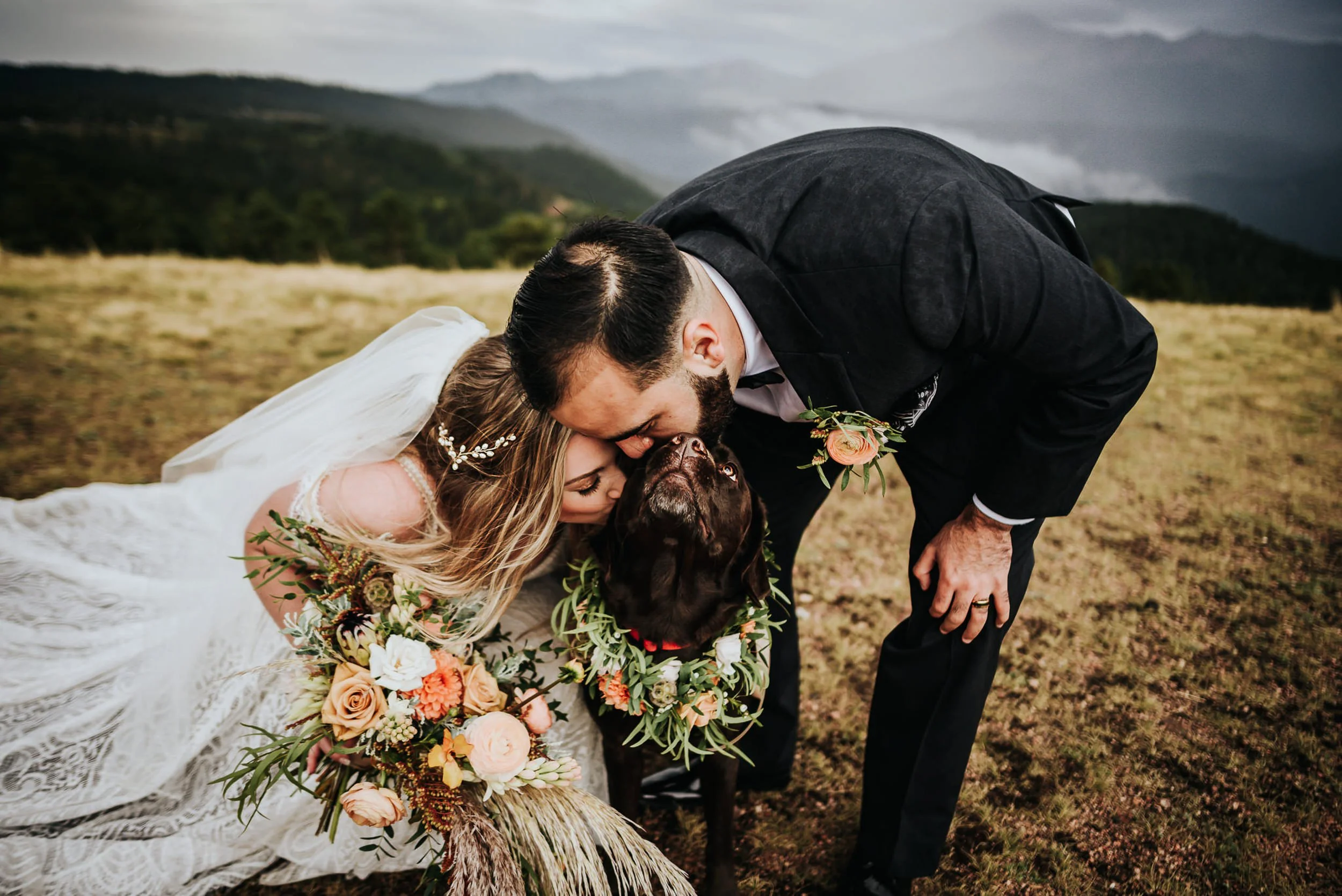 A bride and groom lean down to kiss their dog on the forehead, she holds a flower crown bouquet, mountains behind them.