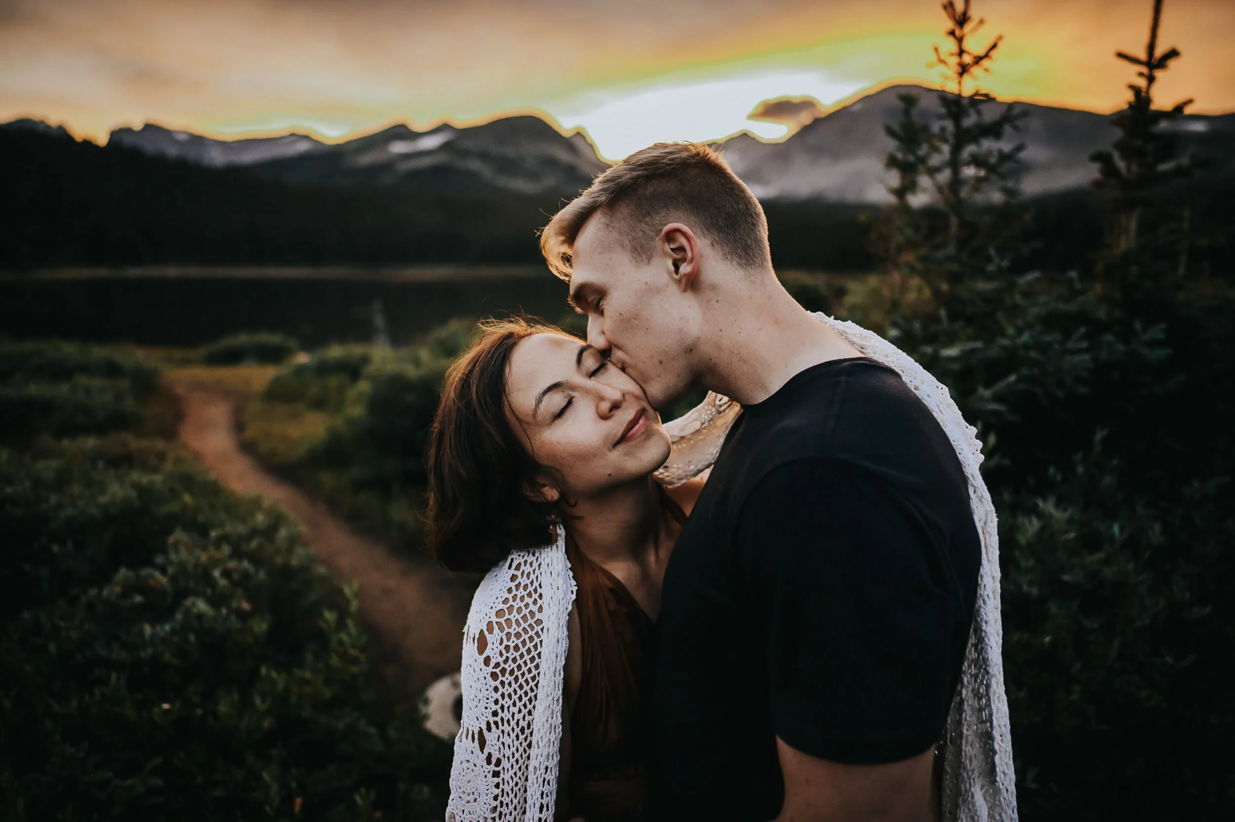 A couple shares a kiss at golden hour with Colorado mountain peaks glowing behind them.