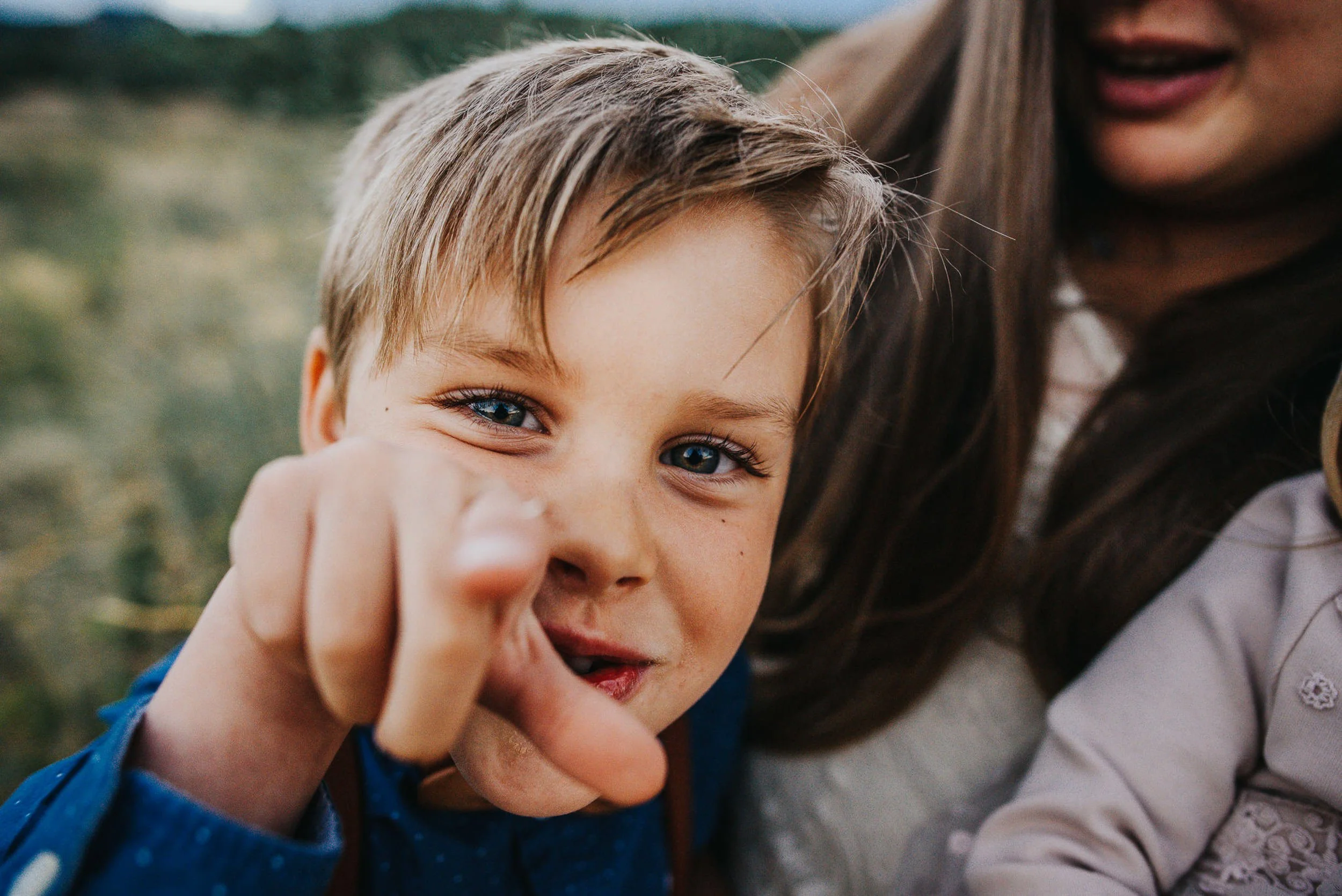 A blond boy with bright blue eyes grins and points directly at the camera.