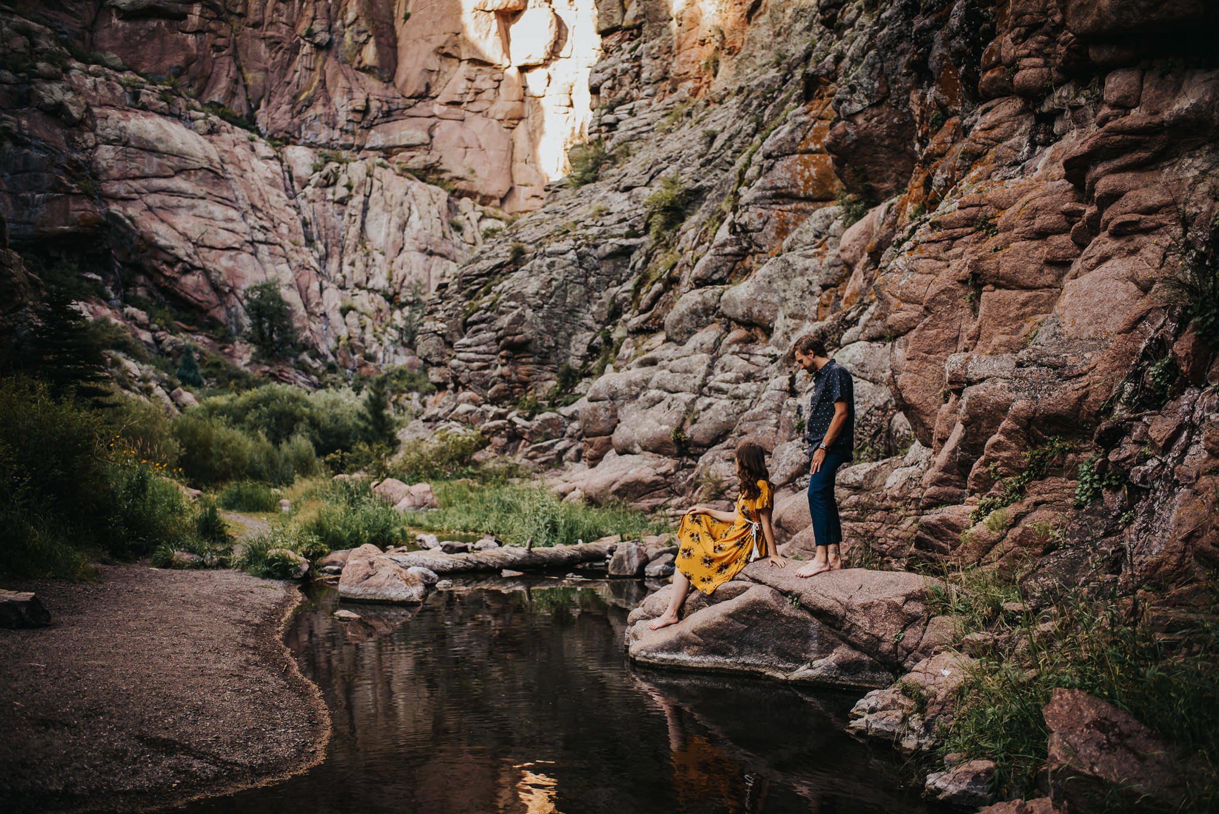 A couple picks their way carefully down a narrow canyon trail together, dramatic rock walls rising around them.