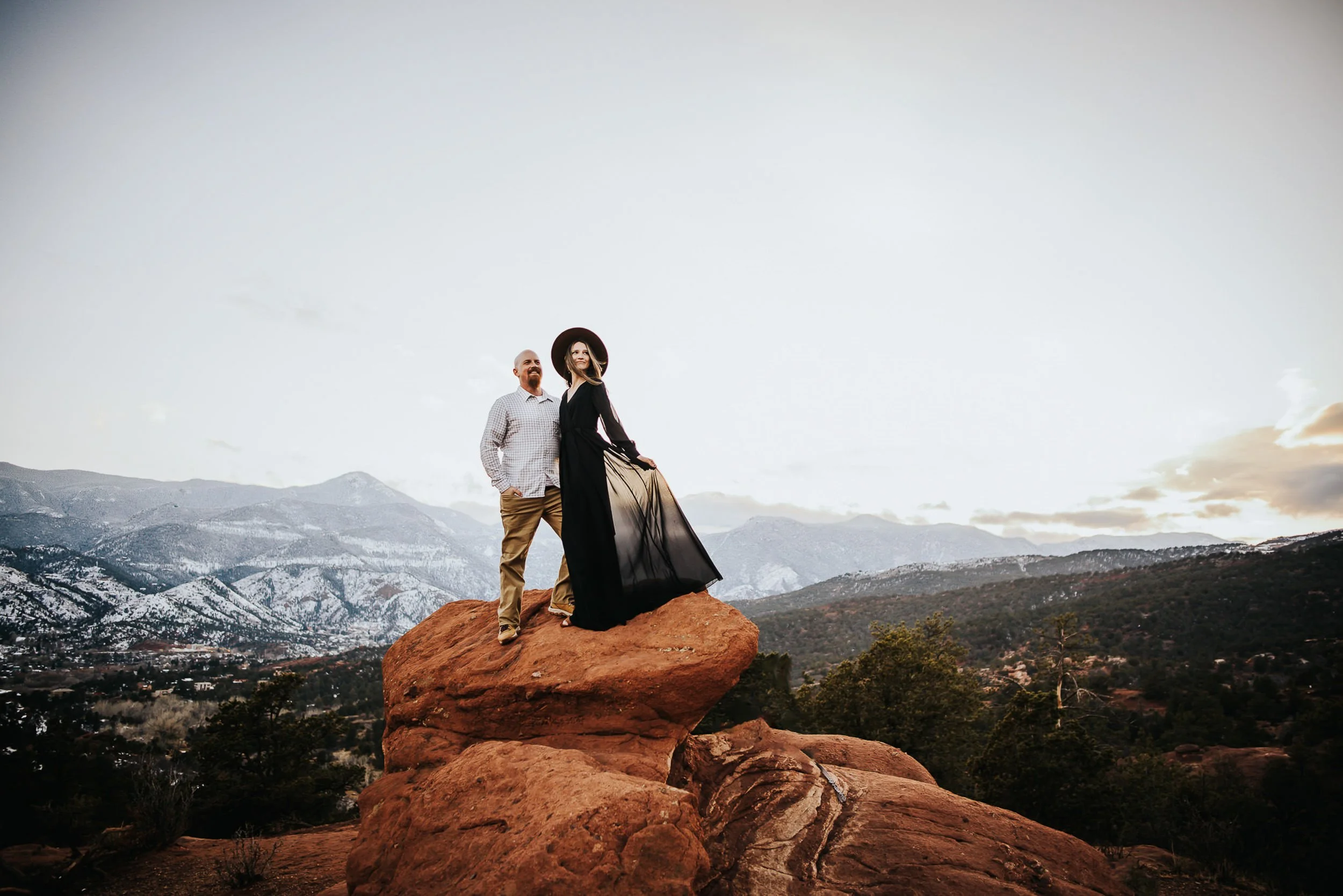 A couples stands tall at Garden of the Gods during a wintery engagement session wearing a black dress and hat.
