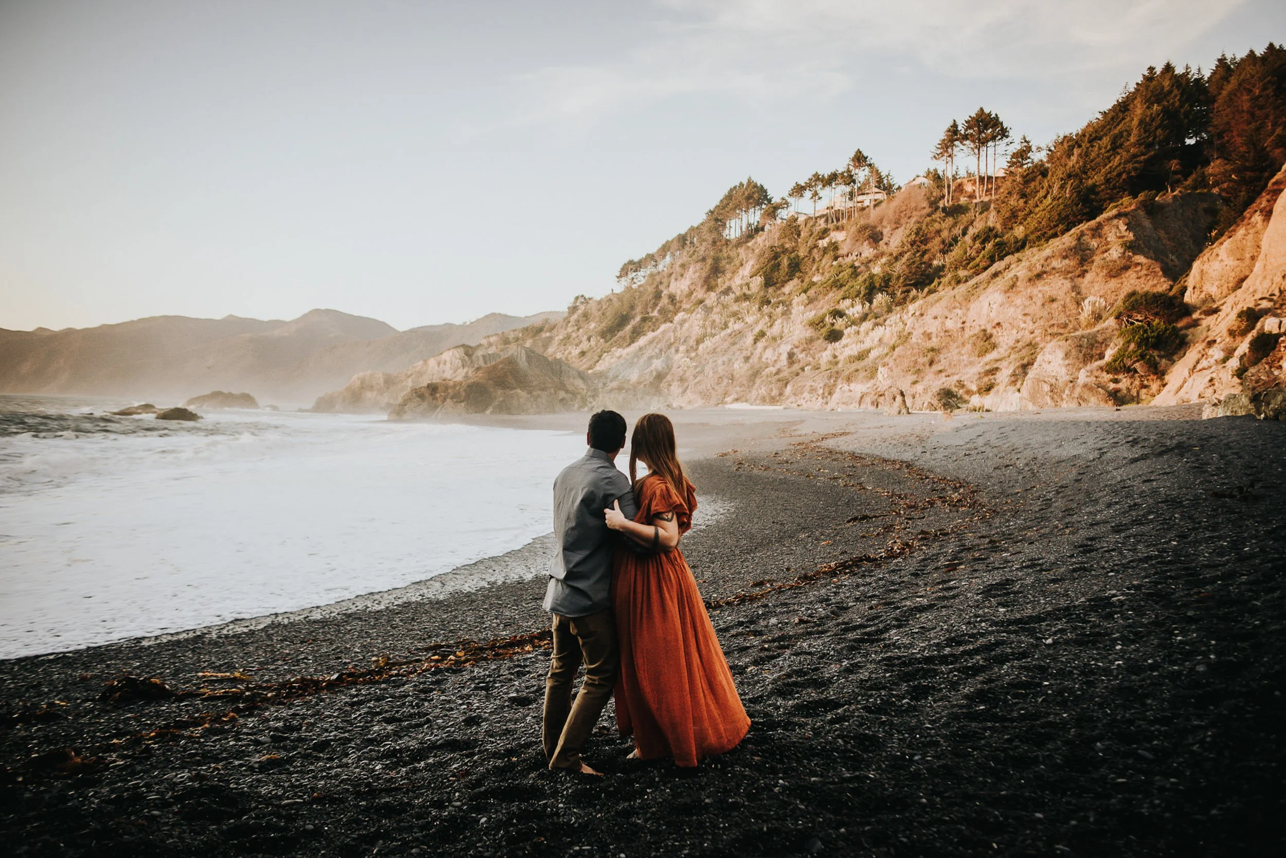 A couple embraces on a dark rocky coastline, she in a rust dress, dramatic coastal landscape behind them.