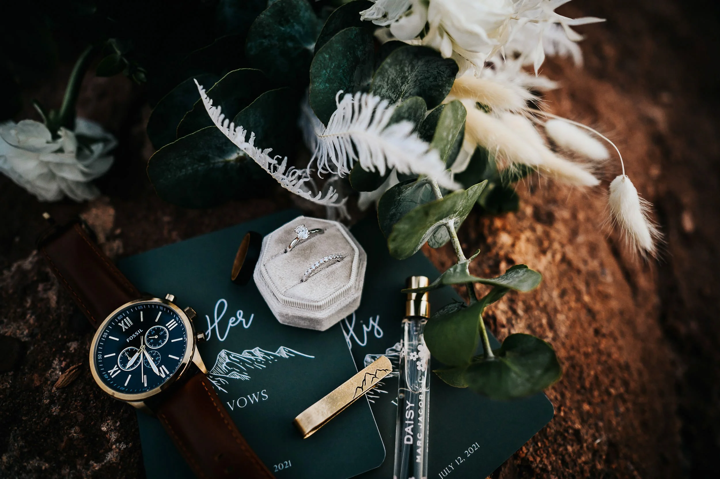 A wedding flatlay with rings, a velvet ring box, a tiara, greenery, and calligraphy details on a dark surface.