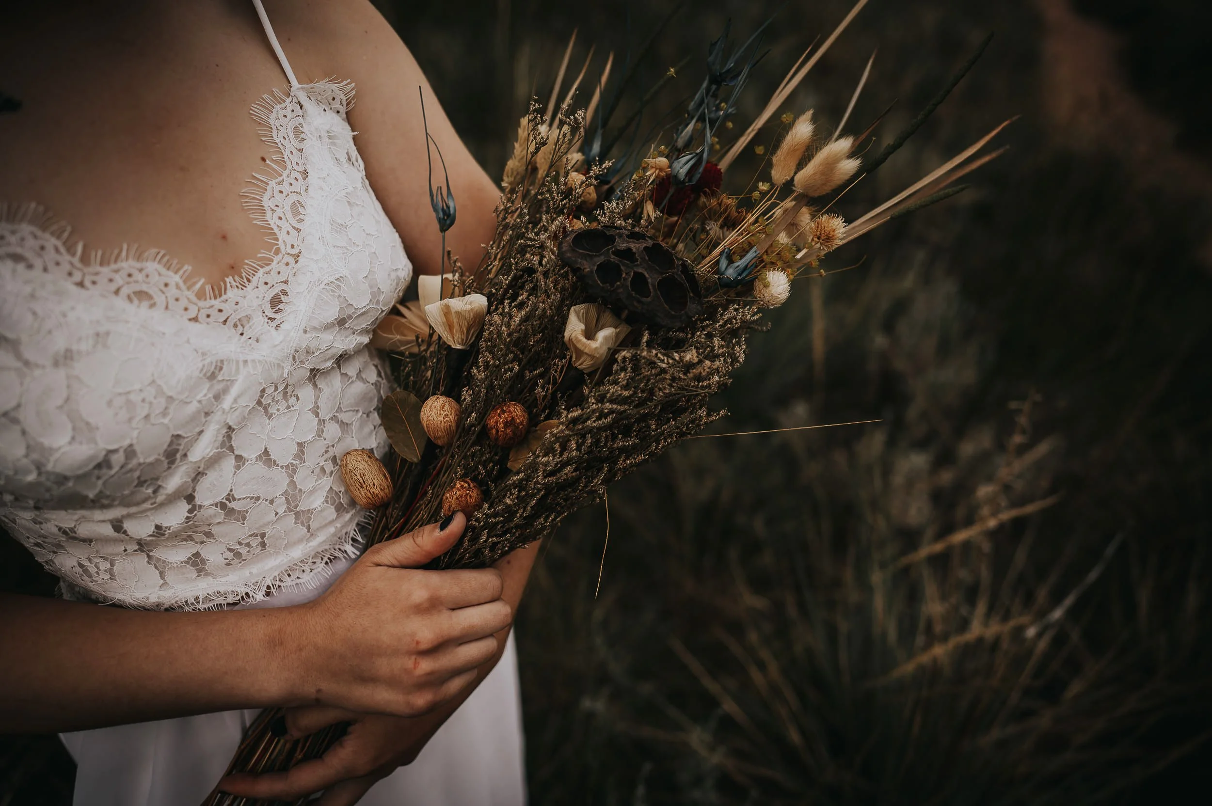 Bride in a lace dress holding a wild dried floral bouquet against a dark natural background.