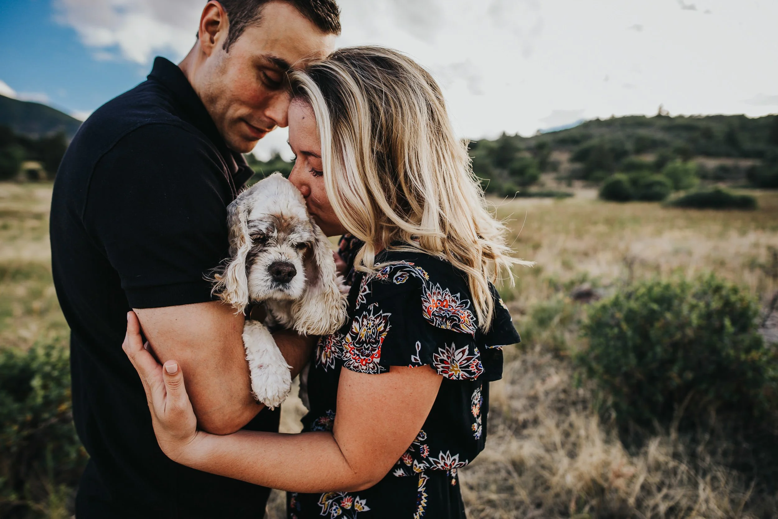 A couple leans forehead to forehead while holding a small fluffy dog between them, Colorado foothills behind them.