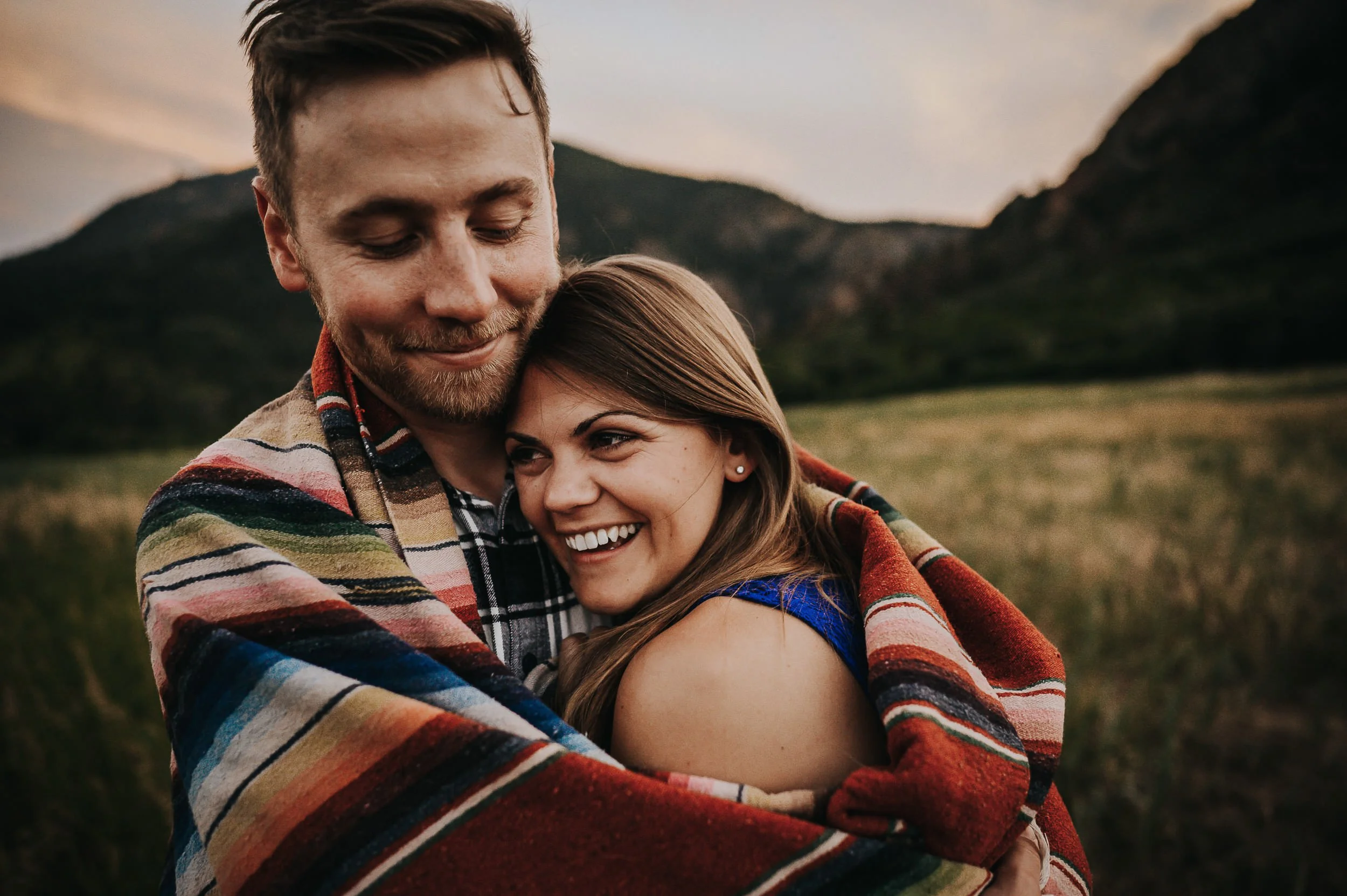 A couple snuggles together wrapped in a blanket, both laughing, Colorado mountains behind them.
