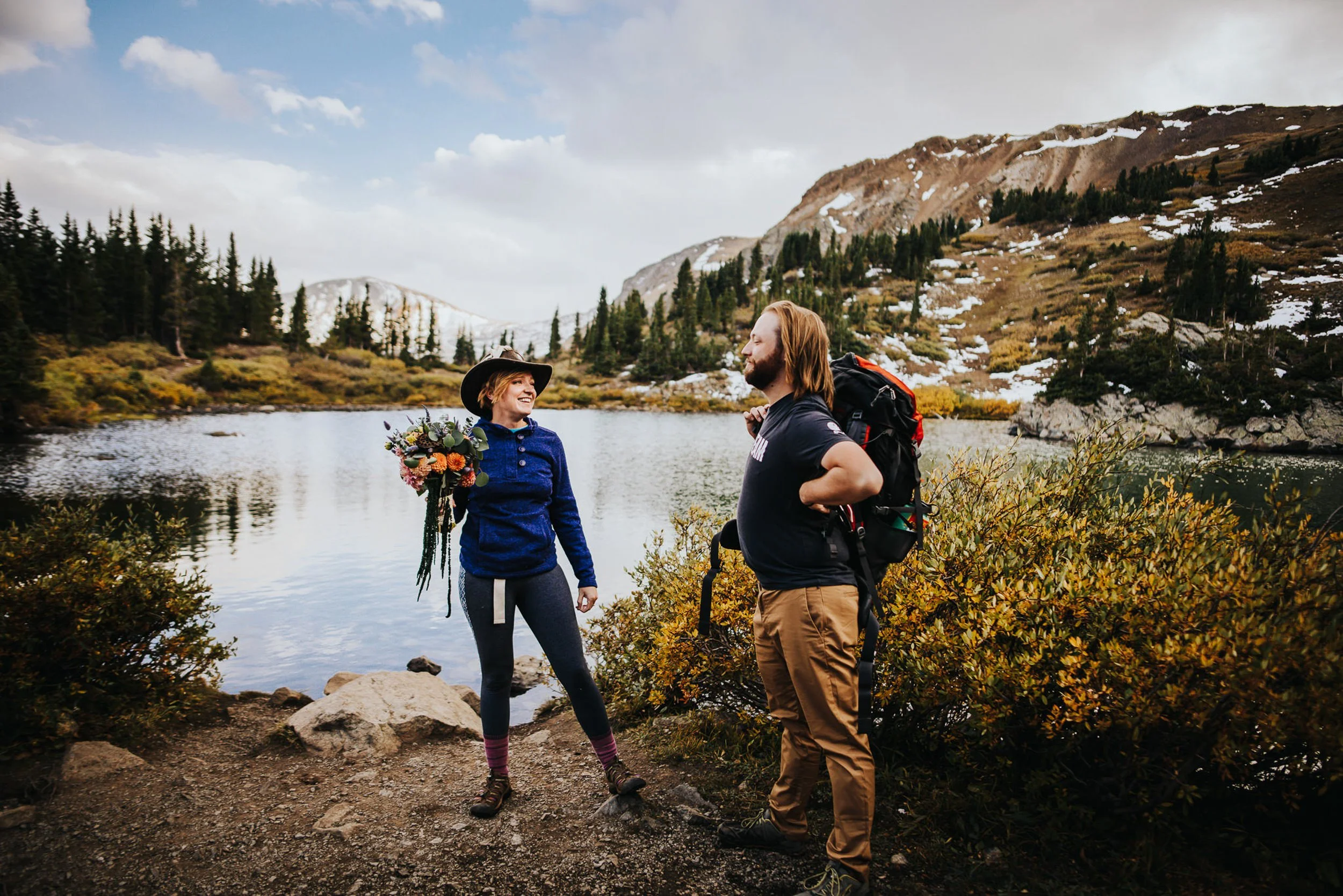 A couple stands together from behind at the edge of a Colorado alpine lake, fall foliage and peaks surrounding them.