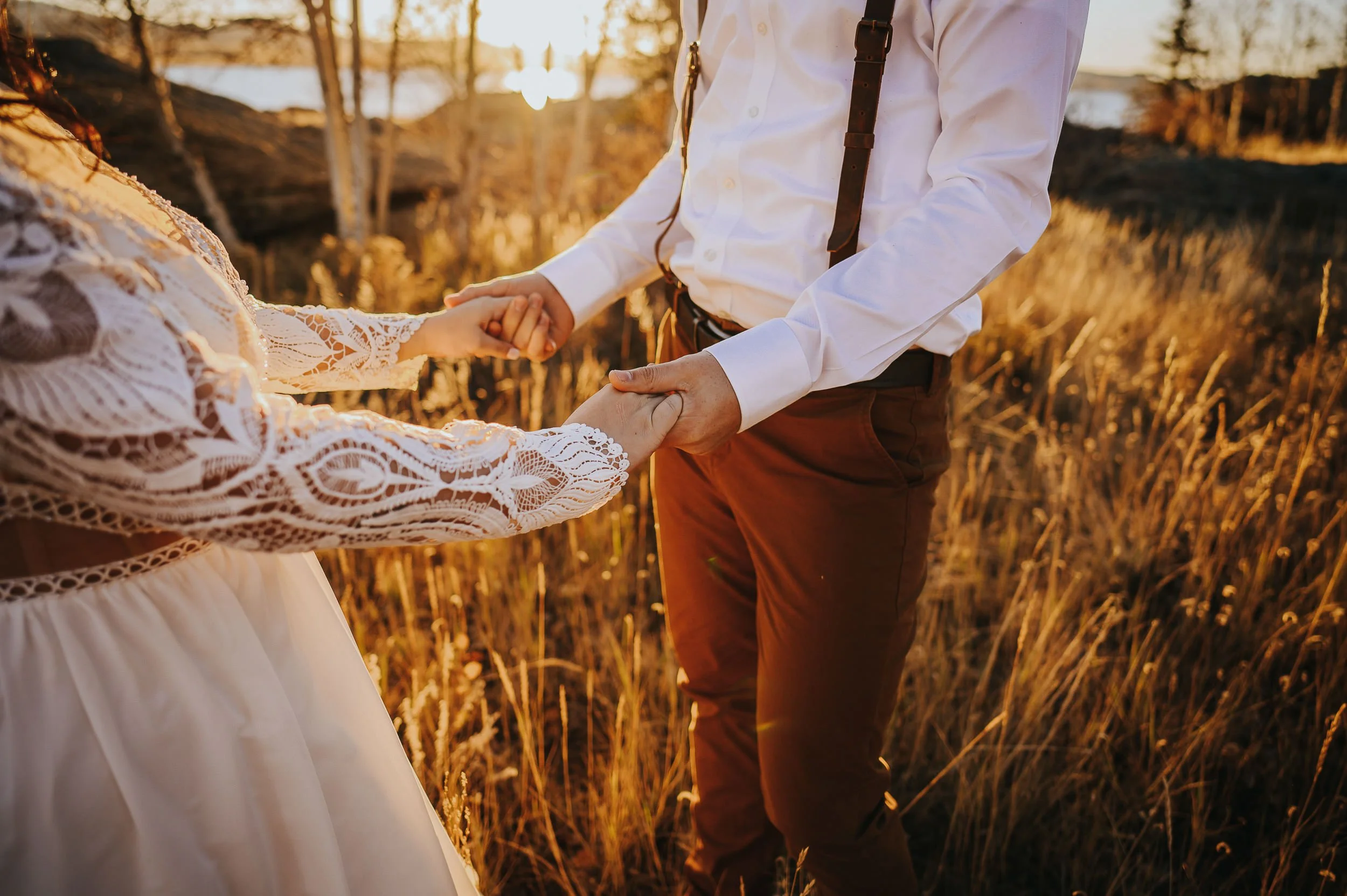 A woman in a boho lace dress holds her partner's hand in a golden Colorado field, her intricate sleeve detail visible.