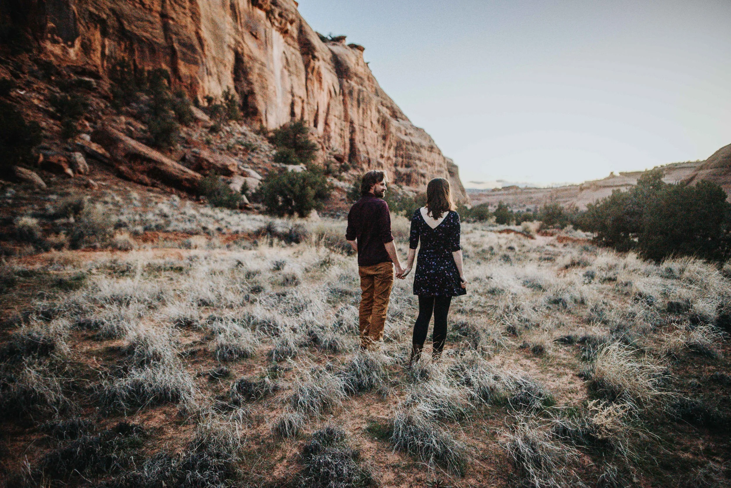 A couple walks hand in hand through a dramatic red rock landscape, wide shot with canyon walls behind them.