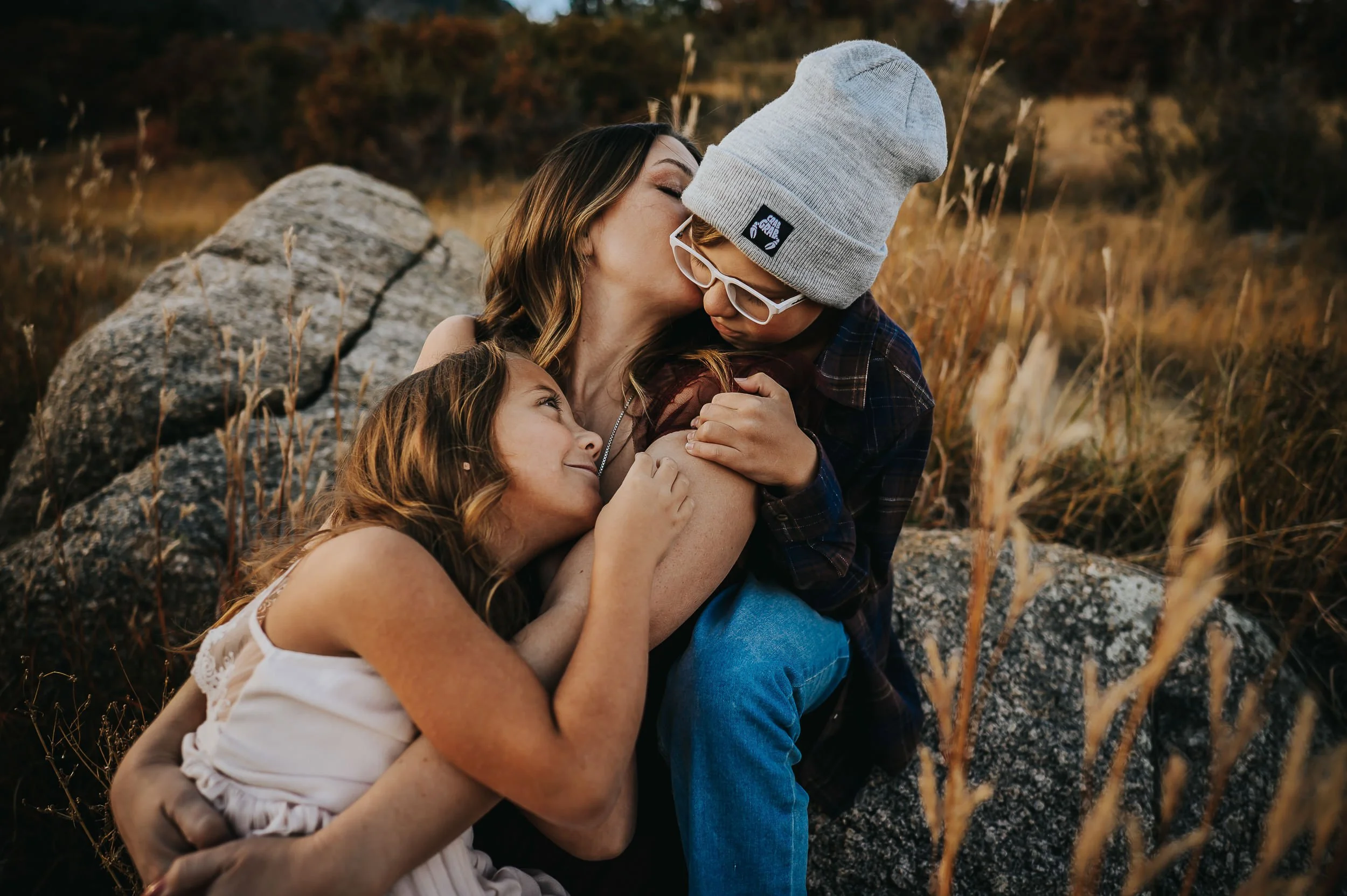 A mom, son, and daughter laugh together on boulders, both in beanies and winter layers.