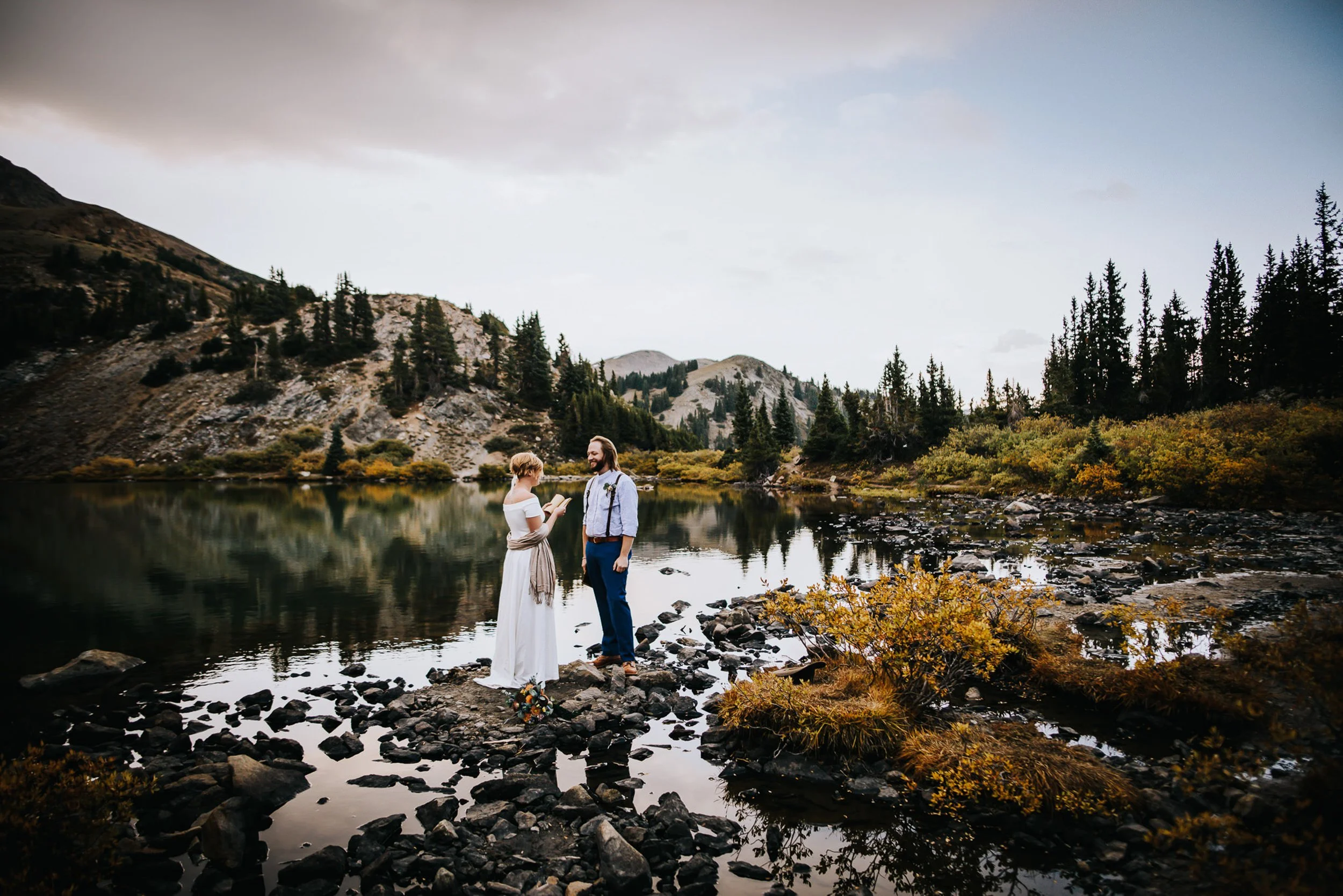 A bride and groom stand at the edge of a still alpine lake, surrounded by fall foliage and peaks.