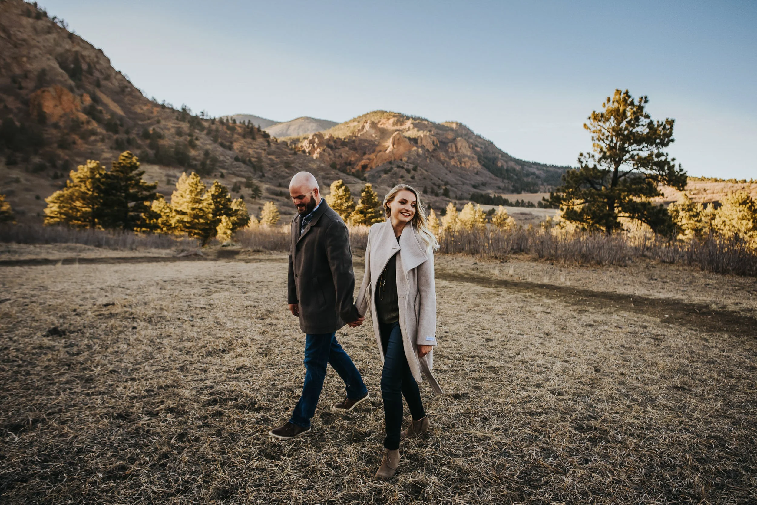 A couple walks hand in hand through a Colorado autumn landscape, mountains and golden foliage behind them.