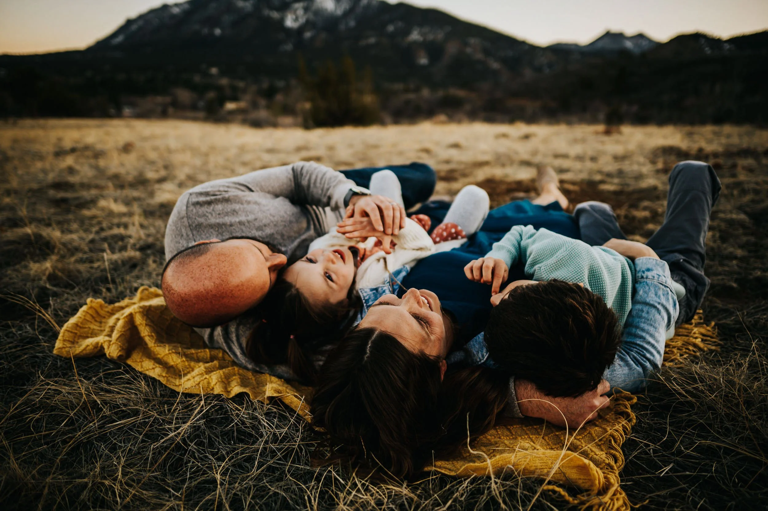 Family of four lay on a yellow blanket in the grass and look up at the sky with mountains in the background.