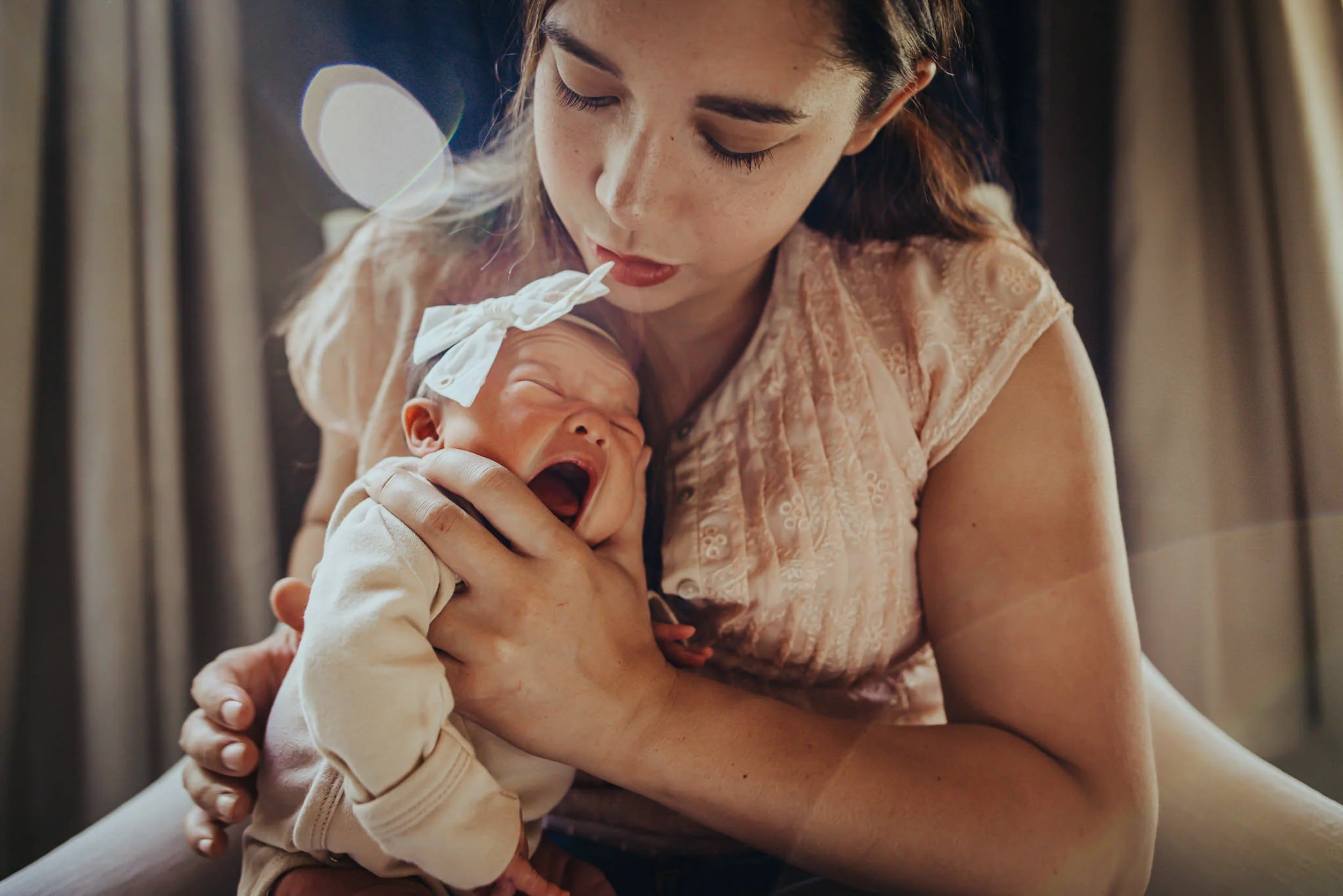 A mother holds her newborn close in soft indoor light, the baby's tiny face visible against her chest.