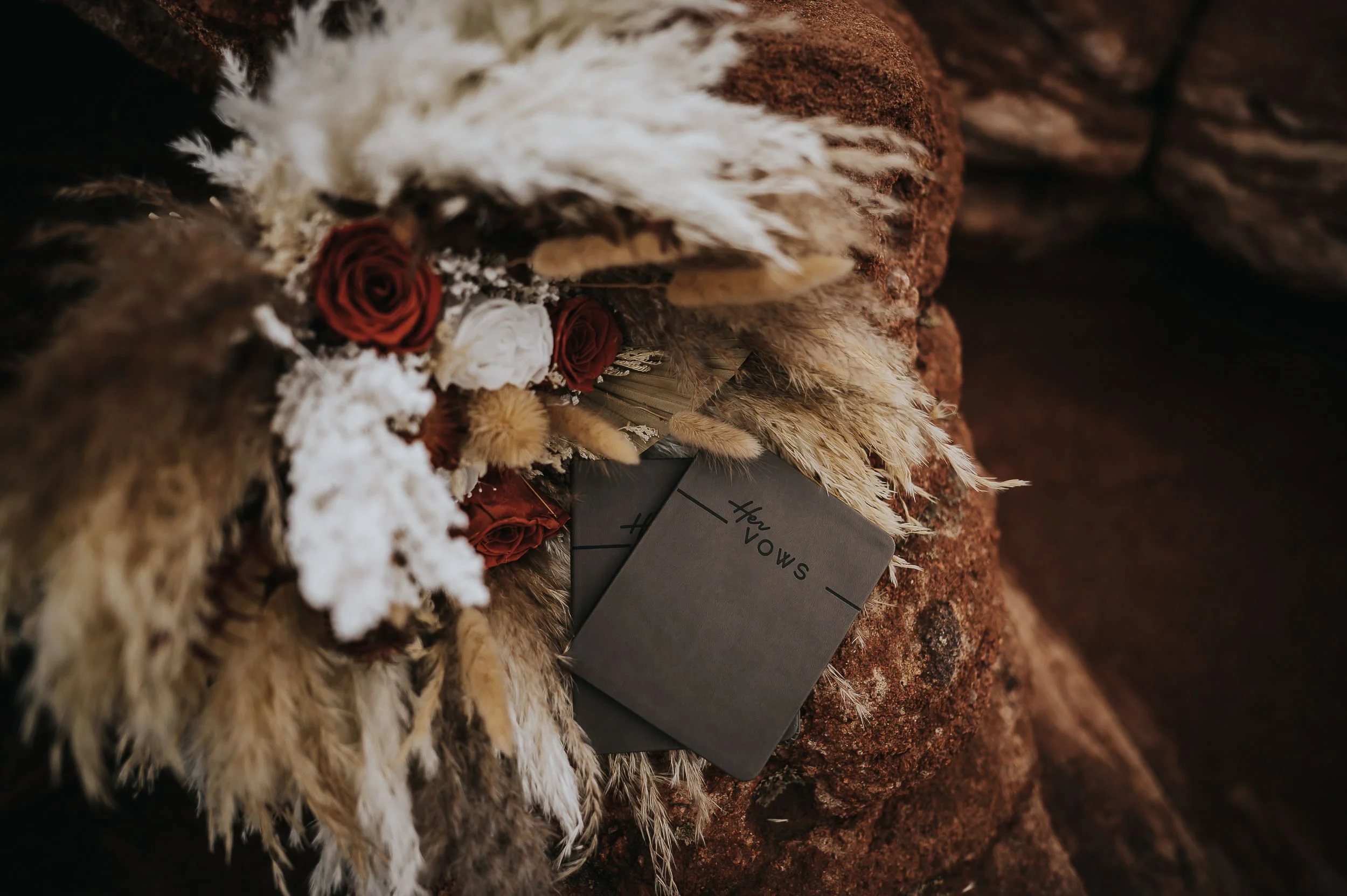 A wedding flatlay with a vow book, dried pampas grass, and a deep red floral arranged together.