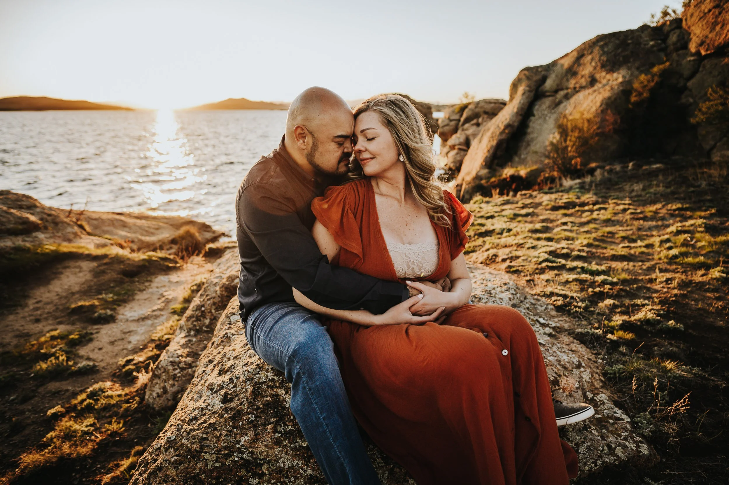 A couple sits close on coastal rocks, he wraps around her from behind, she leans into him in a rust dress.