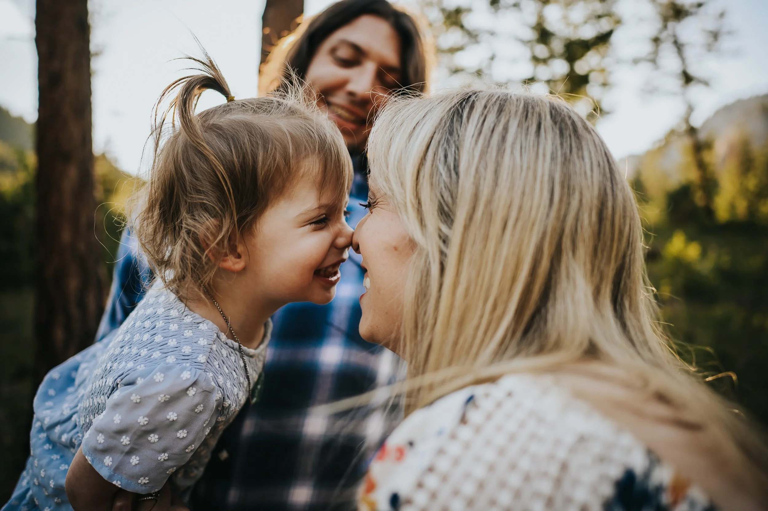 Mother and daughter give each other nose kisses with her father looking on in an evergreen forest.