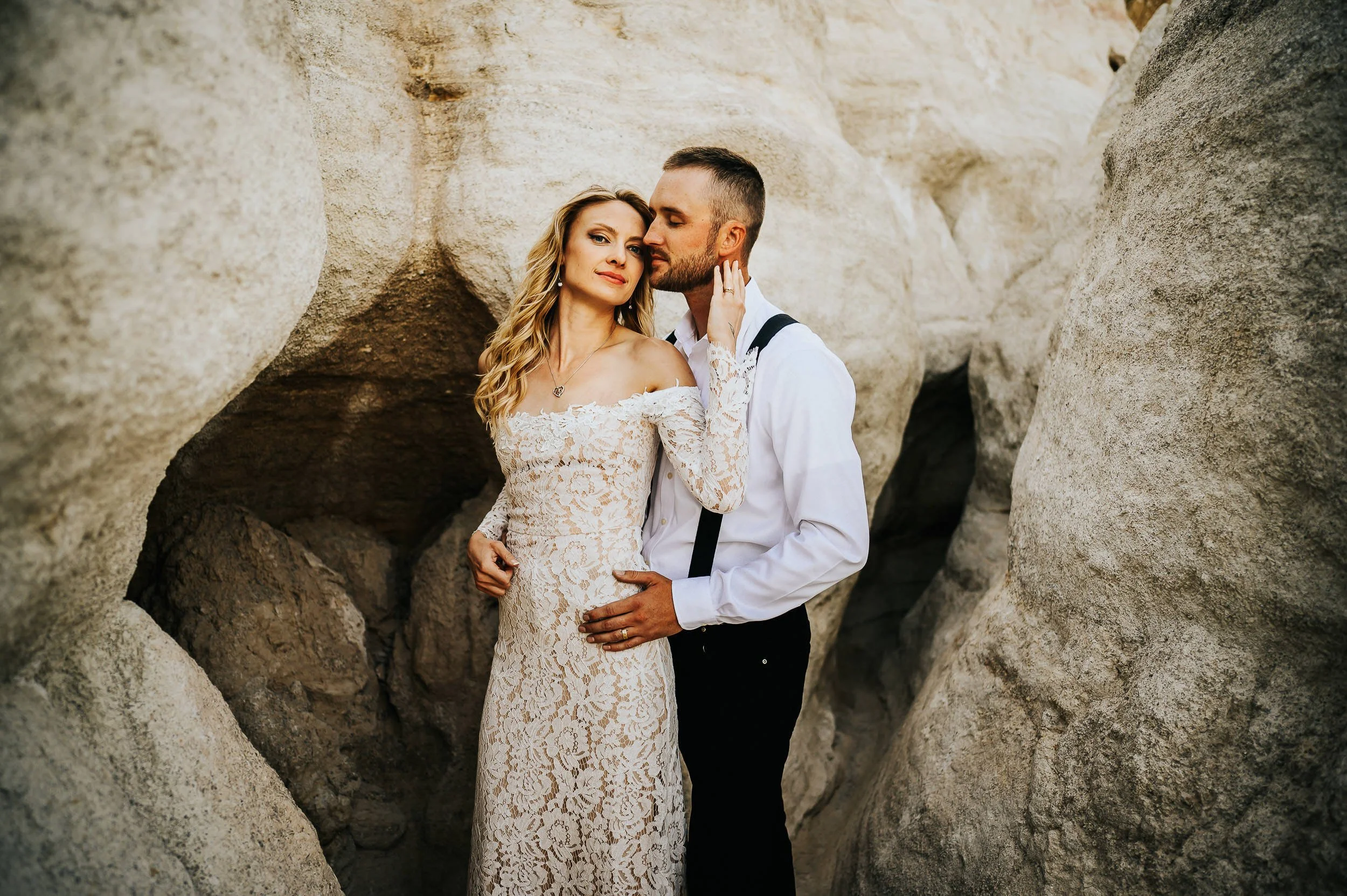 Couple at Garden of the Gods, she wears flowing cream dress, he wears tan pants and light shirt, red rock formations behind them.